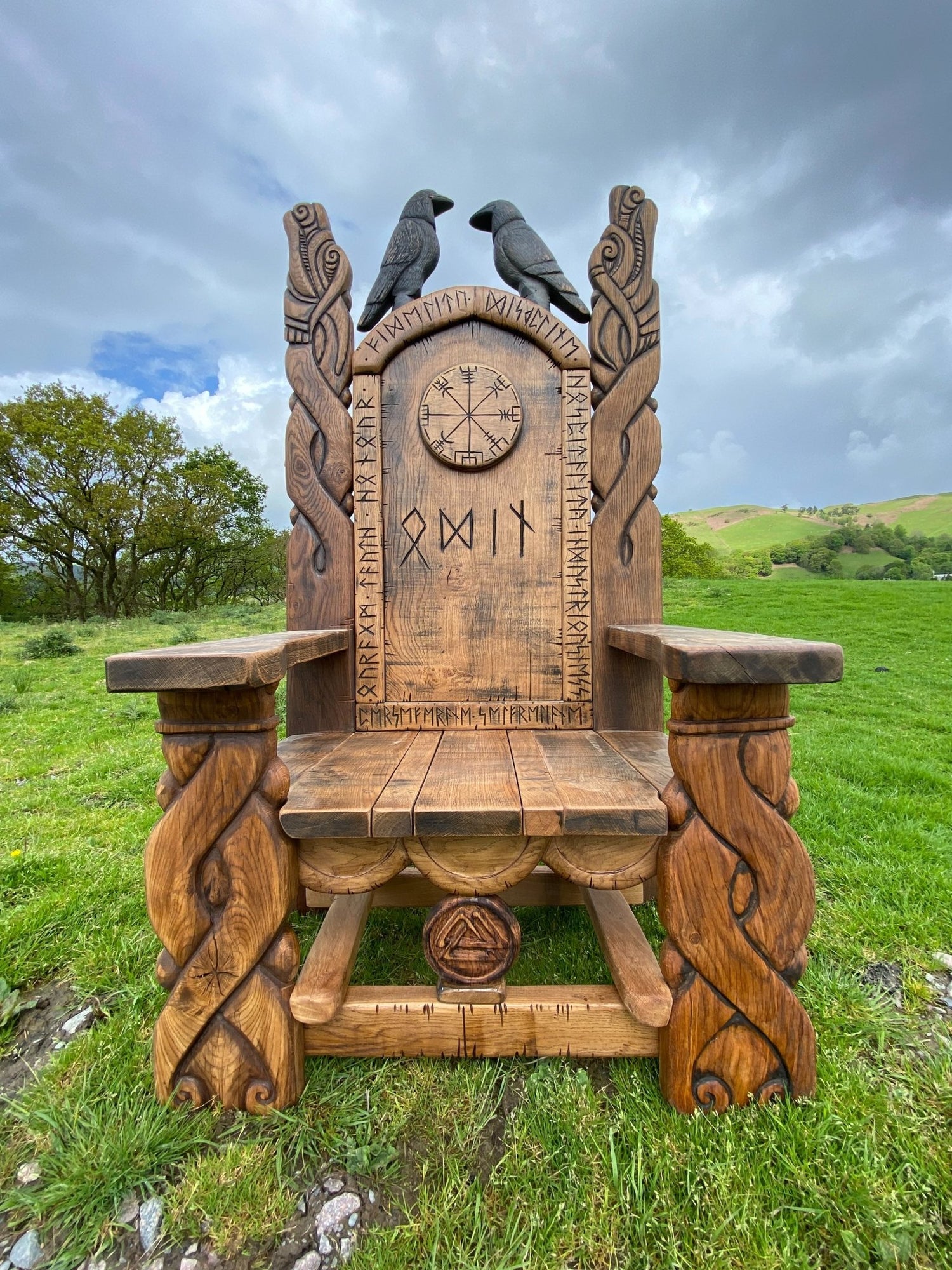 A beautifully handcrafted Viking-inspired wooden carved chair, featuring intricate Norse designs, runes, and two ravens perched on the top of the backrest. Set against a lush green field with a cloudy sky.