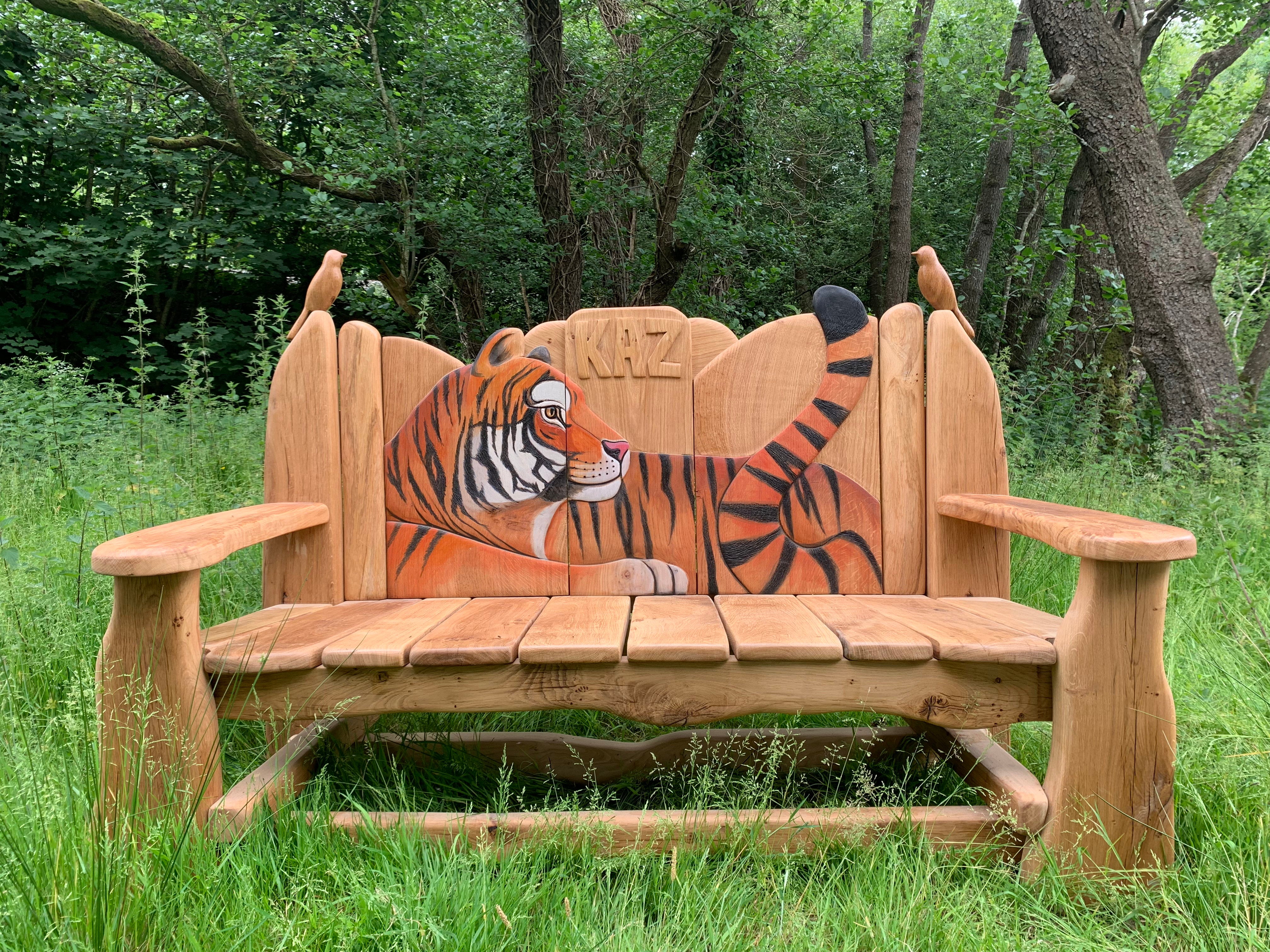 Handcrafted wooden bench featuring a carved and painted tiger reclining on the backrest, with detailed accents of two small bird carvings perched on the sides. Set in a lush, green outdoor environment, showcasing the bench's intricate artistry and natural wood finish