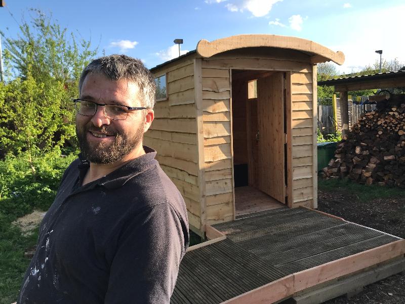 Craftsman standing proudly beside an outdoor composting toilet shed built by Free Range Designs, showcasing sustainable woodworking craftsmanship