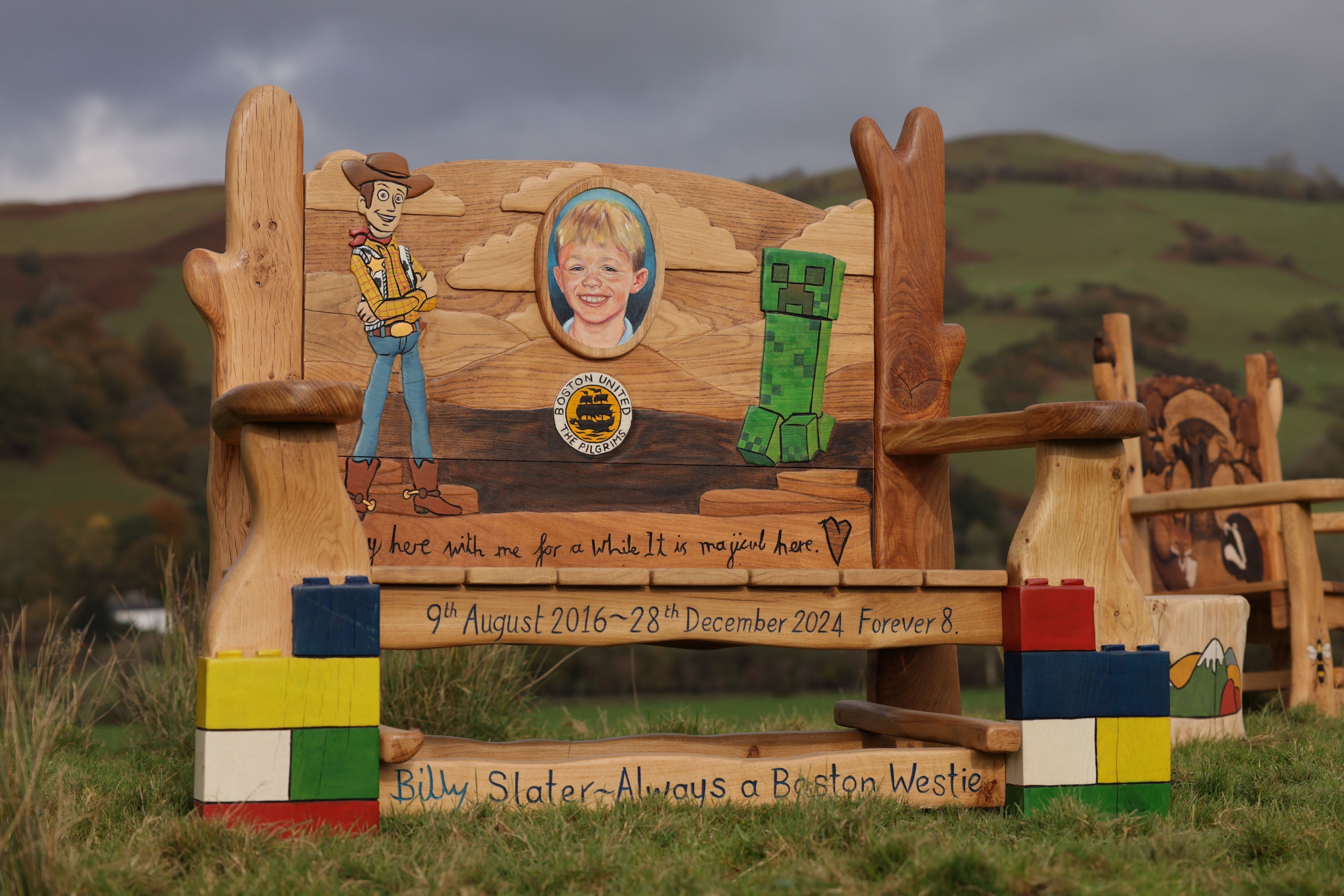 Wooden memorial bench with cowboy figure, child's portrait, and colorful blocks in a natural setting.