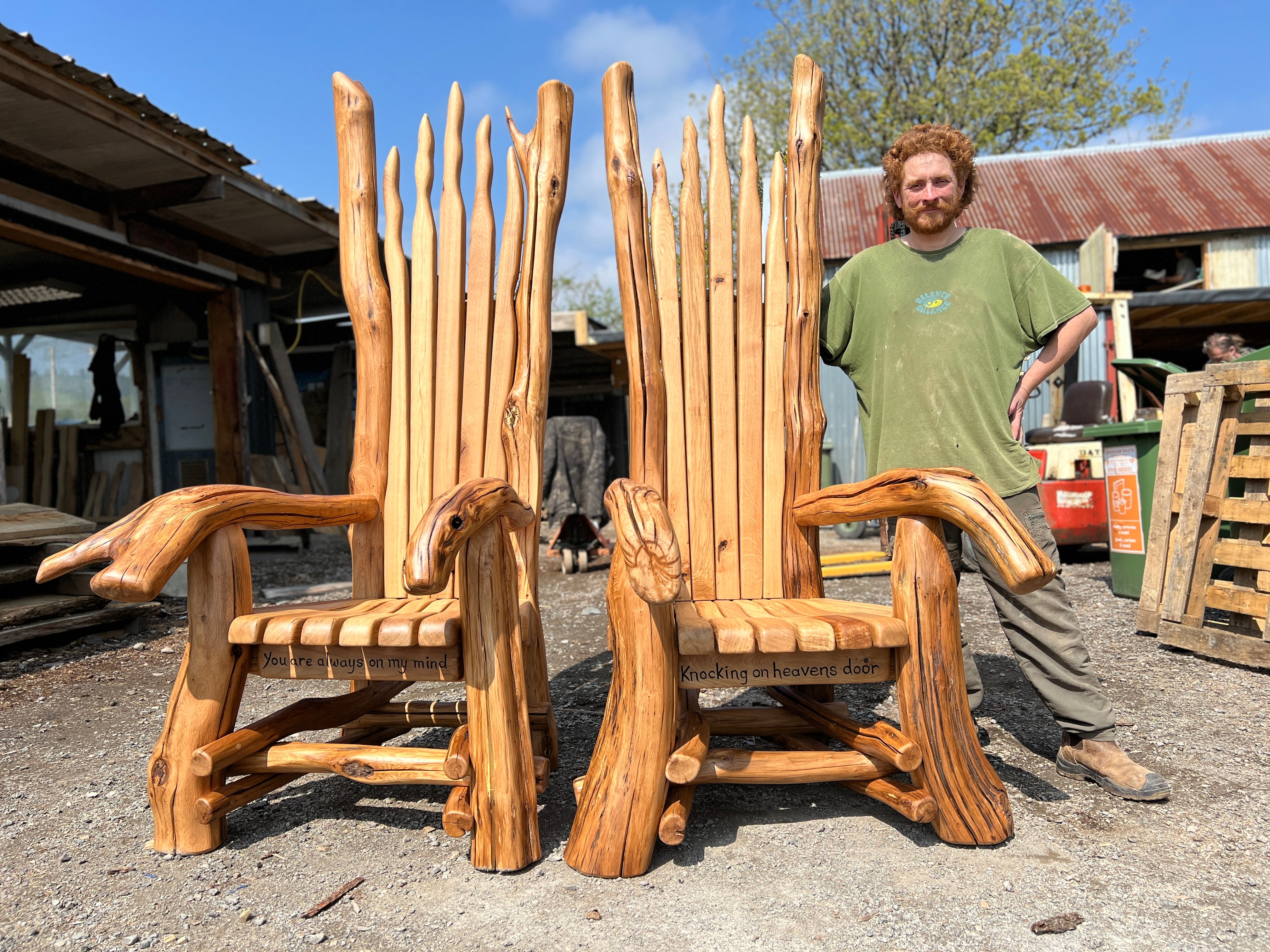 Two tall, handcrafted wooden storytelling chairs with natural, branch-like designs stand side by side in a rustic outdoor workshop. Each chair features expressive animal-shaped armrests—one resembling a bird and the other a seal. The chair on the left has the phrase “You are always on my mind” carved into the seat, and the one on the right reads “Knocking on heaven’s door.” A smiling man in a green T-shirt and work trousers stands proudly beside them.