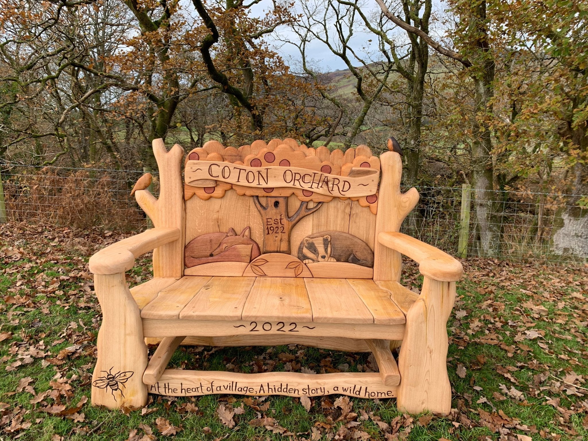 Handcrafted Coton Orchard Bench with intricate wildlife carvings, celebrating local heritage and nature, featuring a fox, bee, and tree motif with engraved inscriptions.