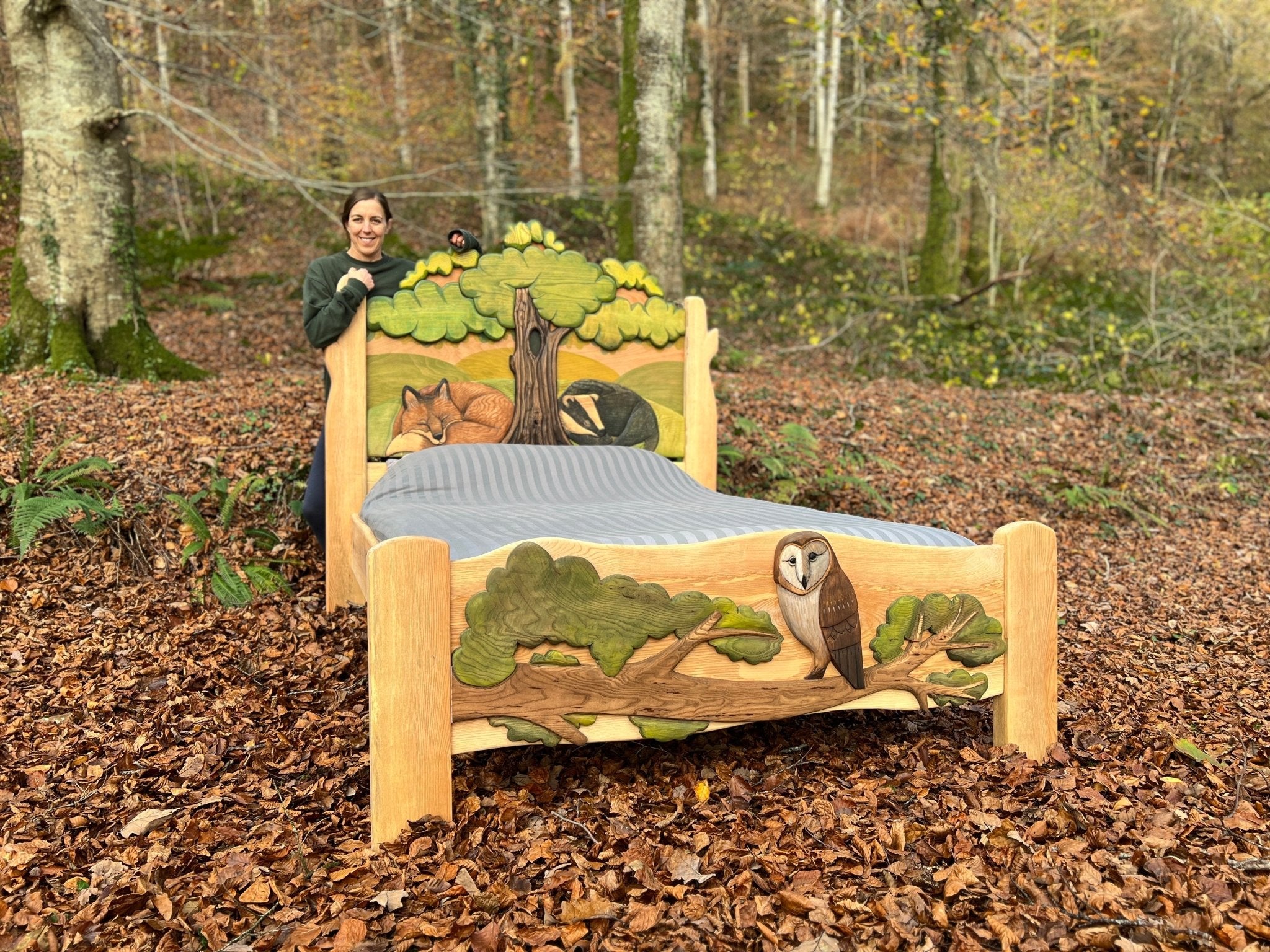 Une femme debout à côté d'un lit en bois sculpté à la main, inspiré par la nature, avec des sculptures détaillées d'un renard, d'un blaireau et d'un hibou, sur fond de forêt. Le lit, fabriqué en bois massif, est le fruit d'un travail artisanal minutieux. Il constitue un mélange unique d'art et de fonctionnalité.