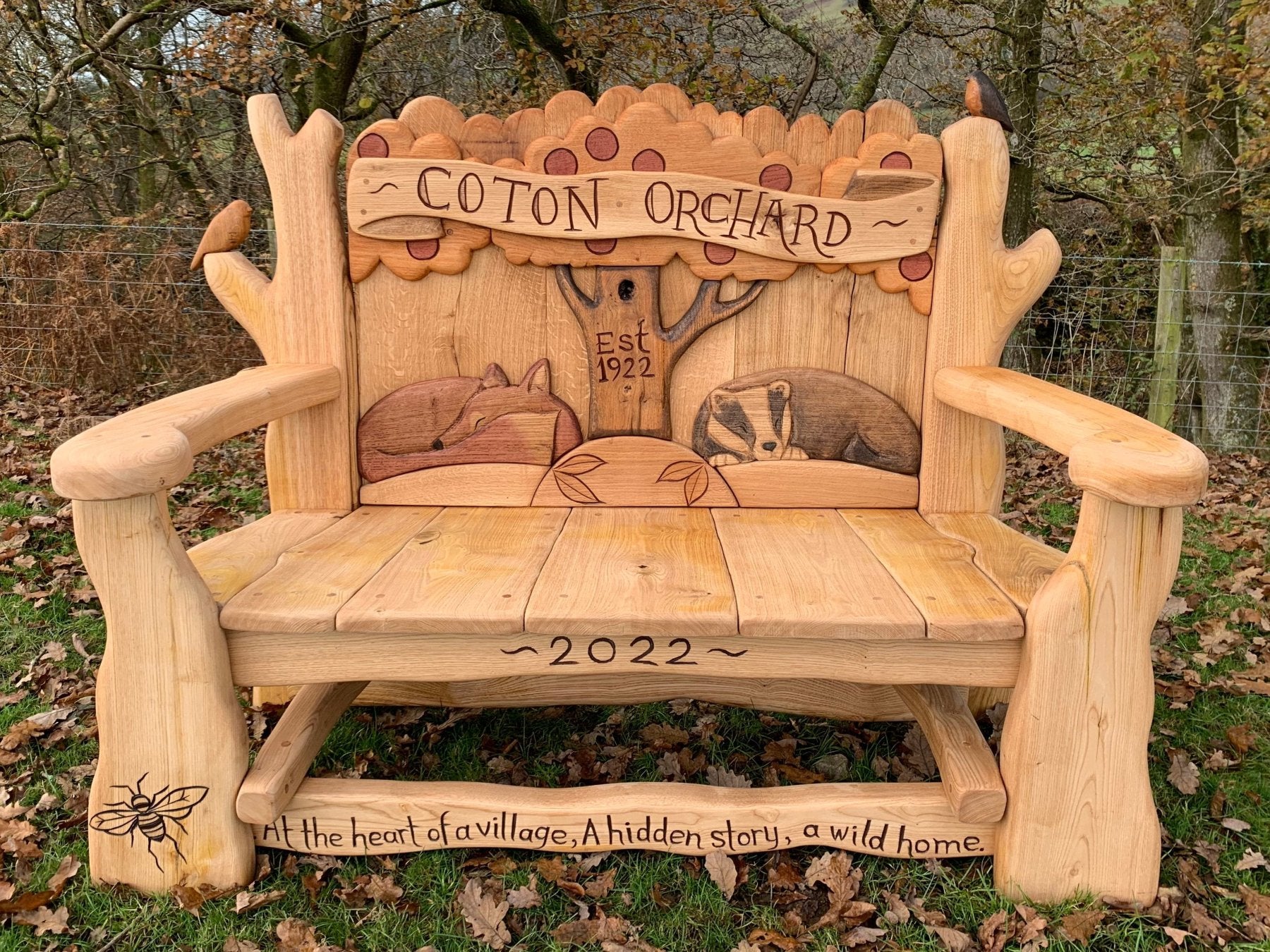 Handcrafted wooden bench with intricate carvings of a tree, fox, and badger, labeled "Coton Orchard Est 1922," featuring a bee and the engraved message "At the heart of a village, A hidden story, a wild home