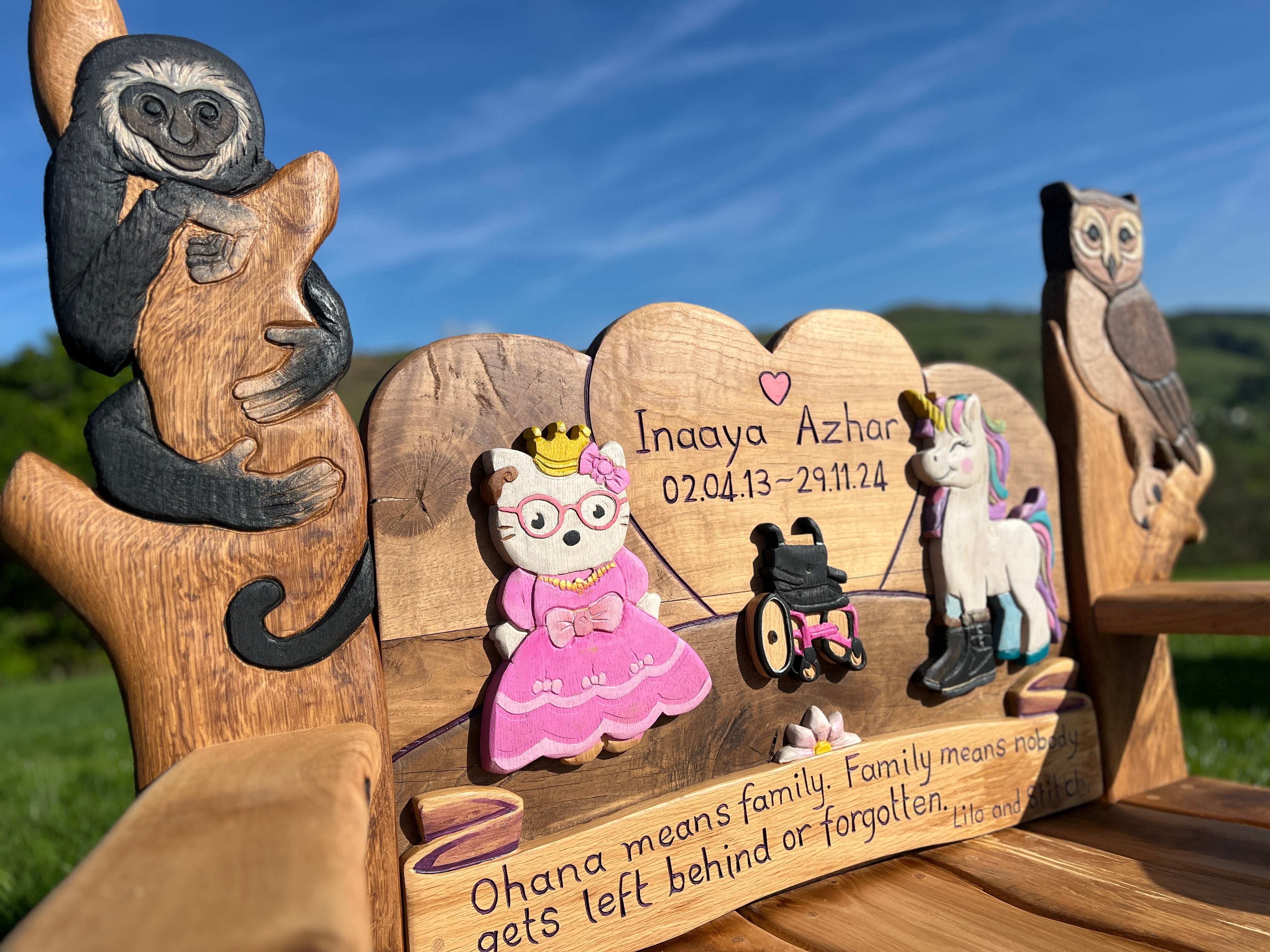 Angled side view of children’s memorial bench featuring carved and painted animals and characters, including a monkey, owl, princess, unicorn, and a personalised engraved tribute to Inaaya Azhar.