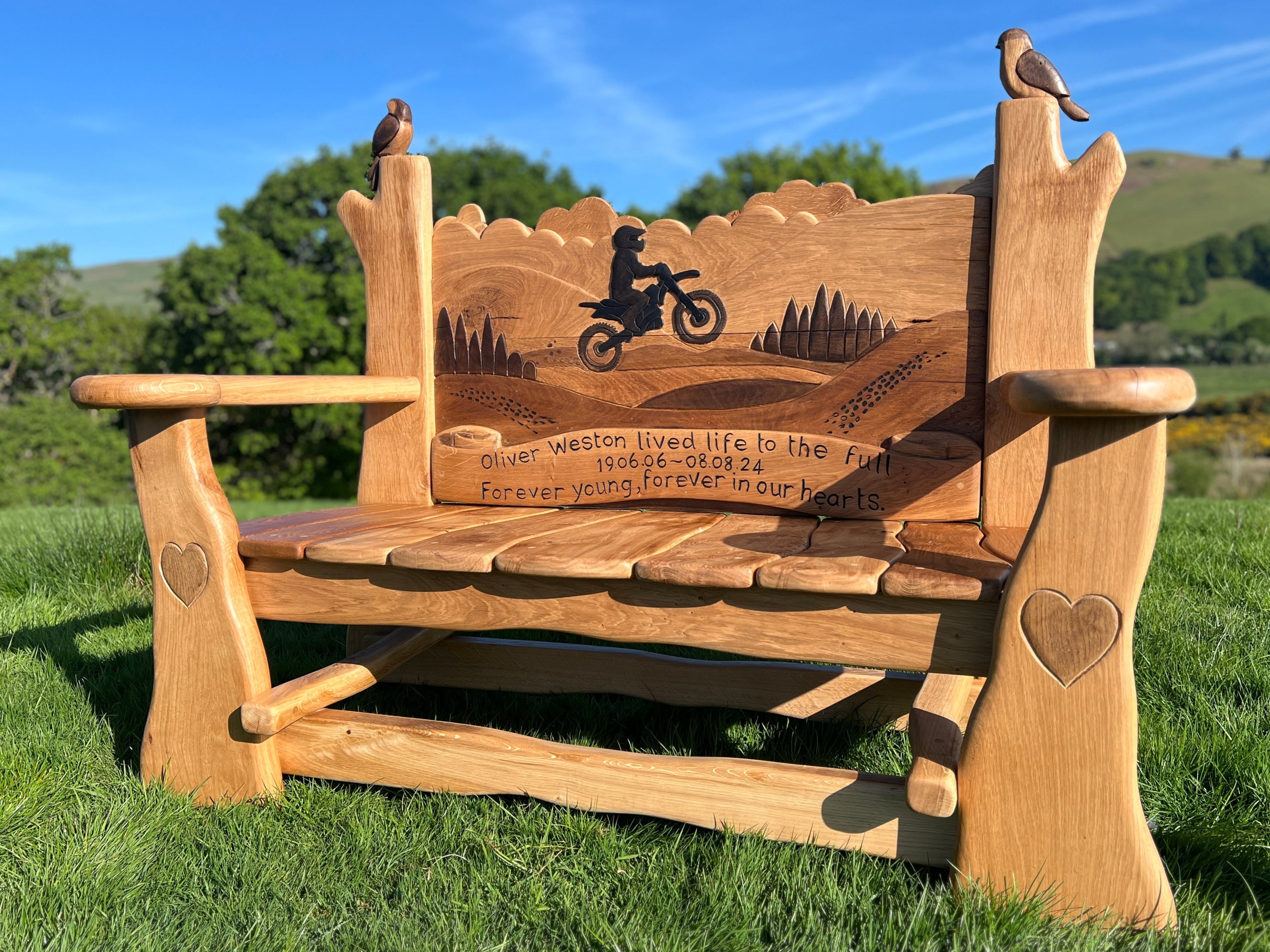 Angled view of hand-carved oak memorial bench featuring a motorbike rider silhouette, engraved dedication to Oliver Weston, heart details on the arms, and carved birds on the back posts.