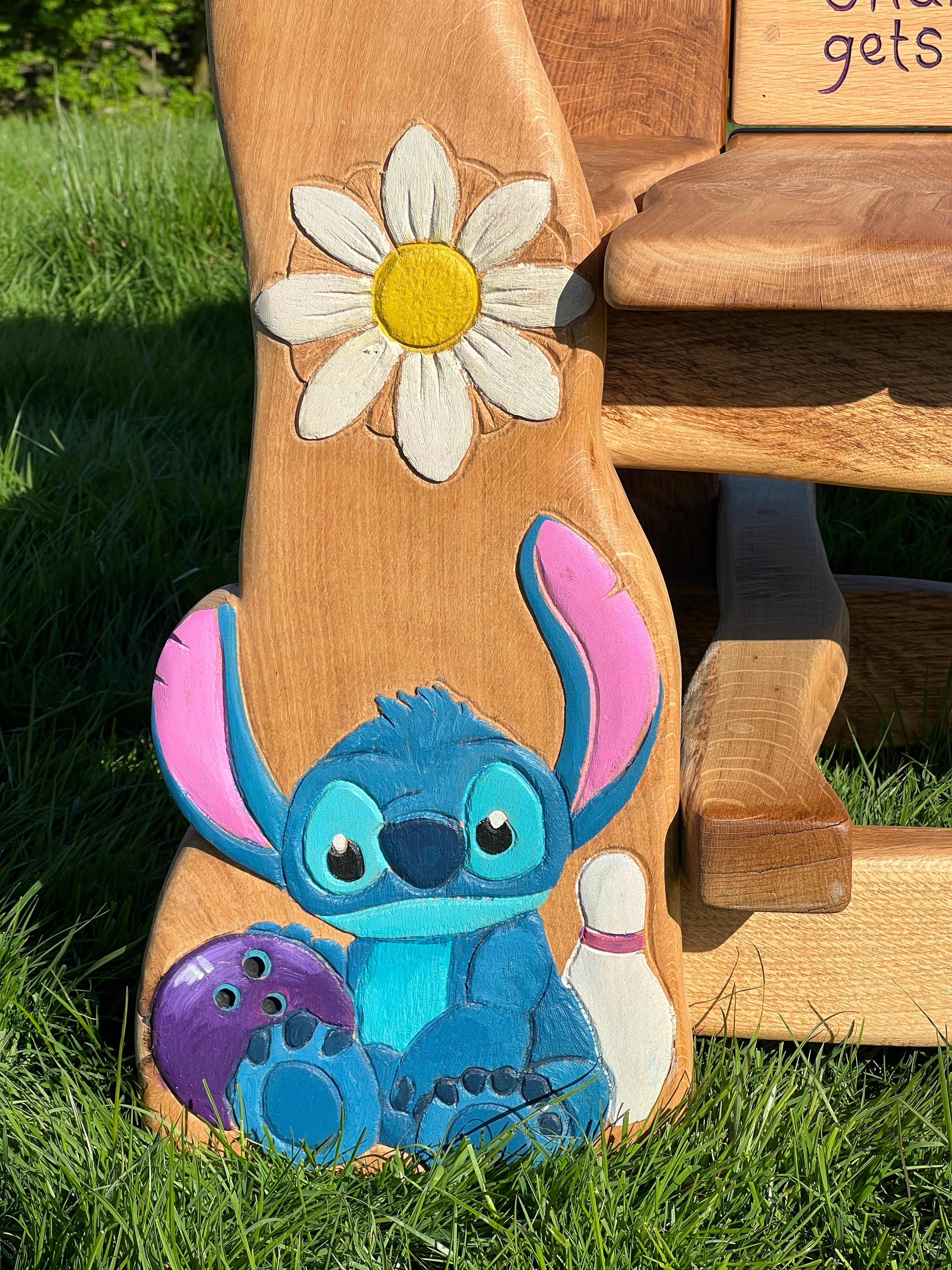 Close-up of hand-painted Stitch character from Lilo & Stitch, holding a purple bowling ball beside a bowling pin, carved into the oak leg of a children’s memorial bench.