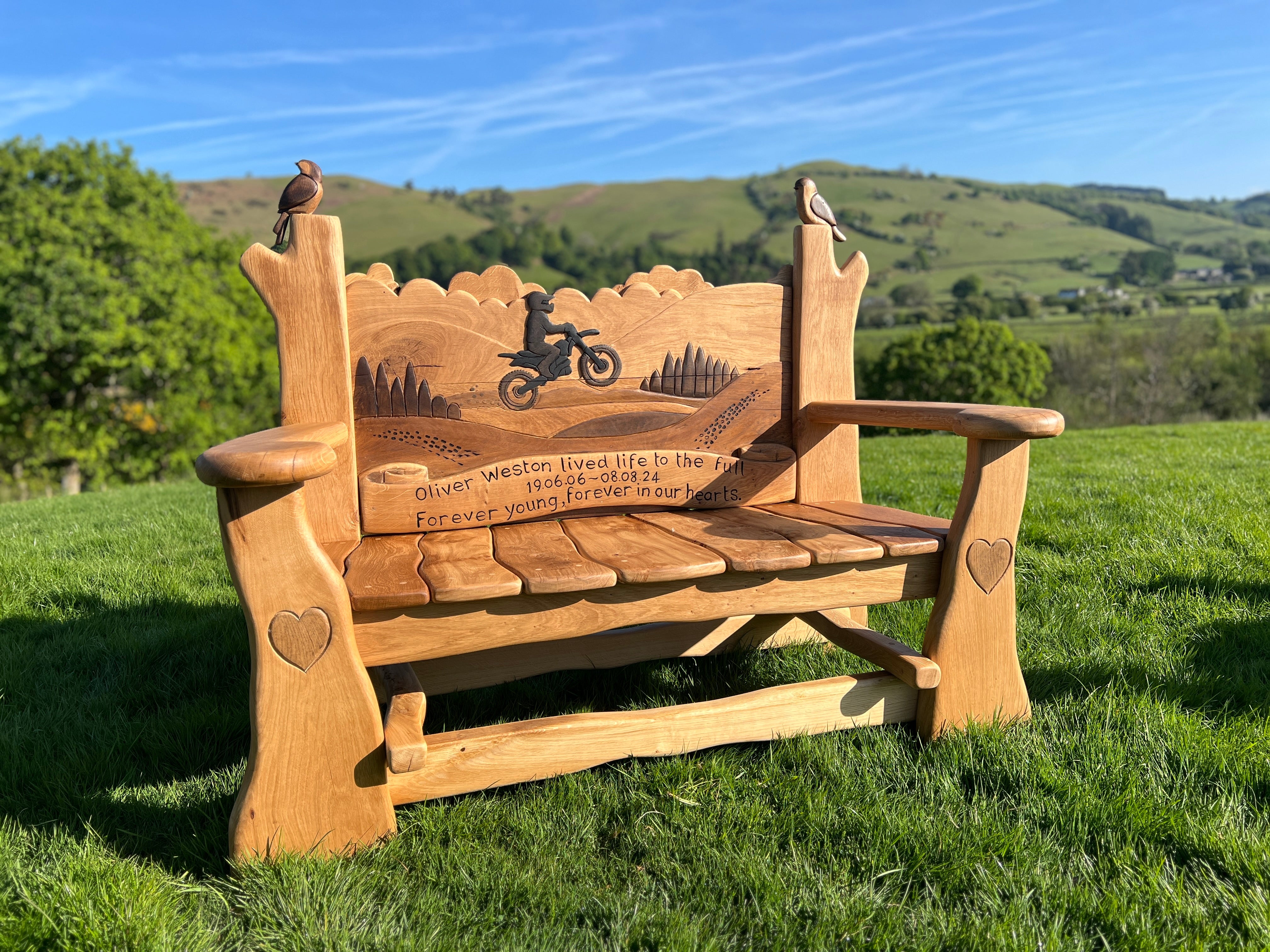 Hand-carved oak memorial bench with motorbike rider silhouette and engraved tribute to Oliver Weston, set in a grassy landscape with countryside background.