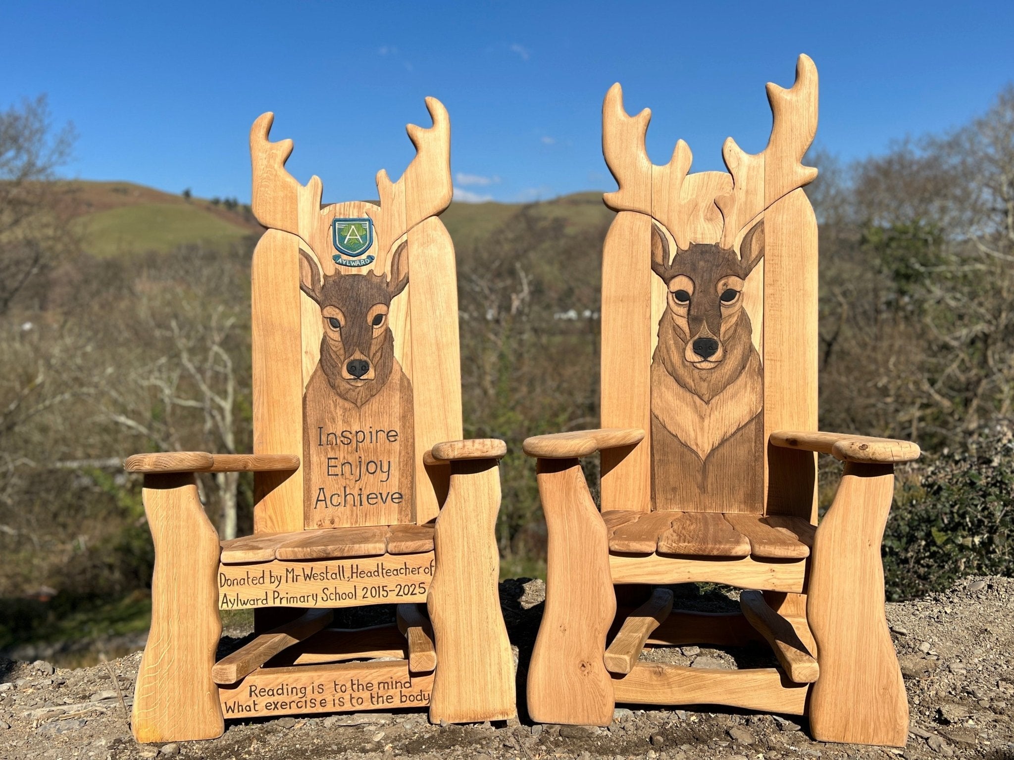 Two handcrafted wooden storytelling chairs featuring carved and painted deer heads with large antlers, set outdoors against a backdrop of rolling hills and leafless trees. The left chair includes a school crest and the words “Inspire, Enjoy, Achieve” along with a dedication to Mr. Westall, headteacher of Aylward Primary School 2015–2025, and the quote “Reading is to the mind what exercise is to the body” engraved on the lower panel.