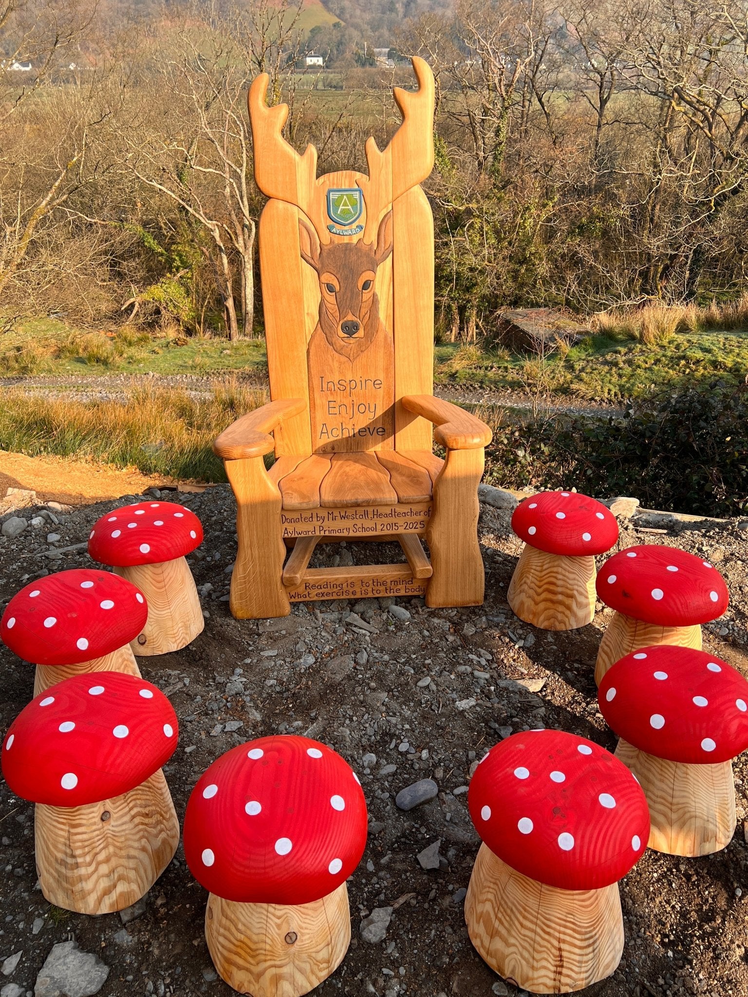 Handcrafted wooden storytelling chair featuring a carved stag and school motto, surrounded by playful red and white mushroom stools, set in a natural outdoor landscape with hills and trees in the background.