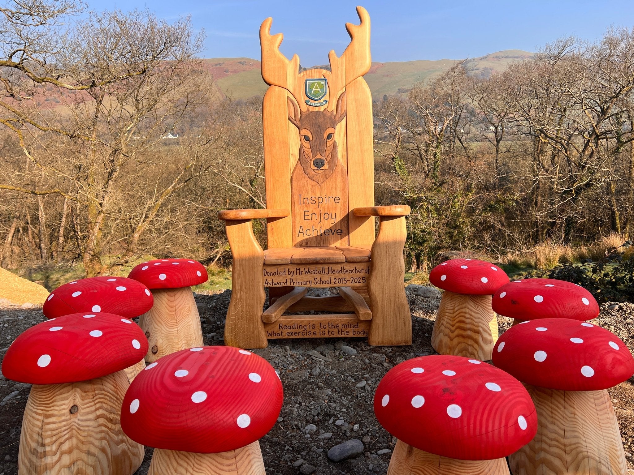 A large wooden storytelling chair with a carved deer design and engraved inspirational messages stands outdoors, surrounded by a circle of whimsical red and white spotted mushroom-shaped wooden stools. The scene is set against a backdrop of bare trees and rolling hills under a clear blue sky.