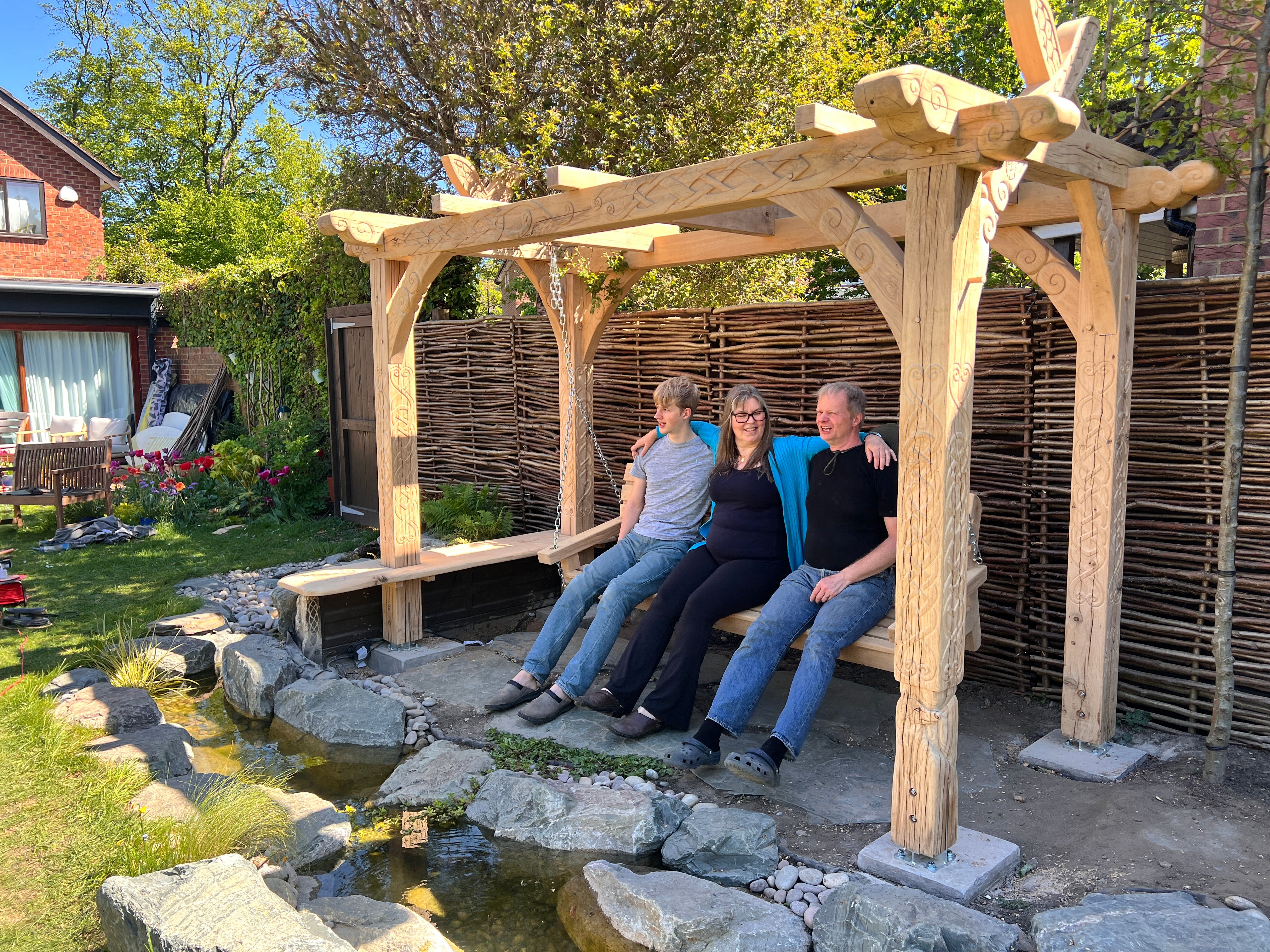 Family seated on the swing bench of the hand-carved Viking oak garden arbour, beside a garden stream in London