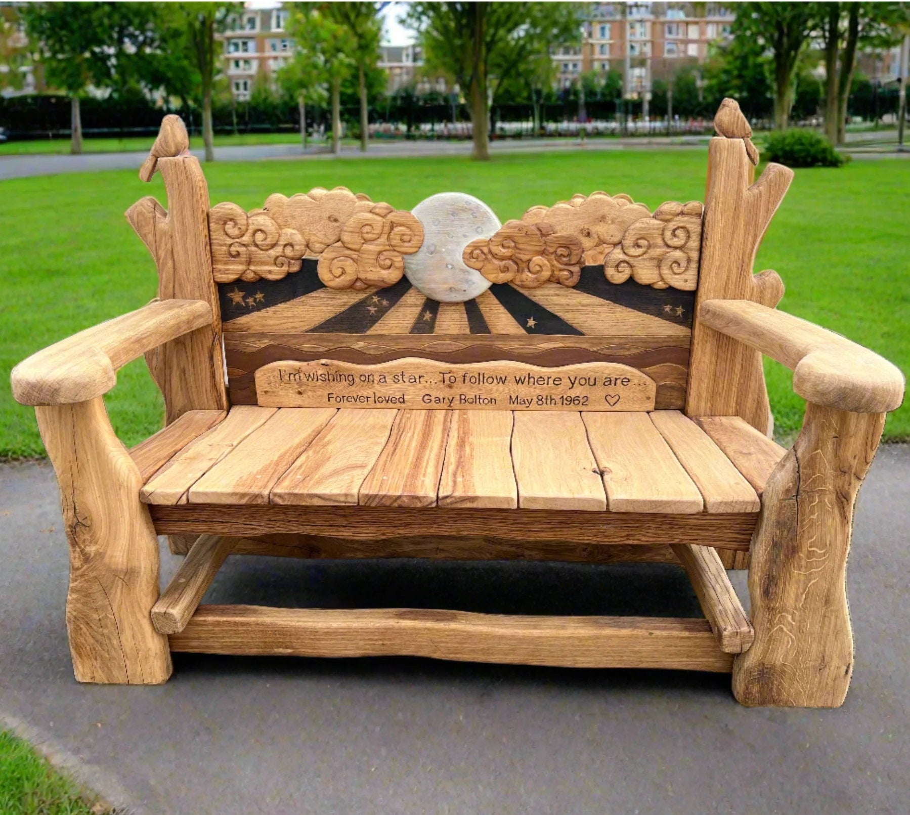 Beautifully crafted wooden memorial bench dedicated to Gary Bolton, featuring a celestial design with carved clouds, a glowing moon, and stars. The inscription reads: 'I’m wishing on a star... To follow where you are. Forever loved Gary Bolton, May 8th, 1962.' The bench is adorned with bird carvings on each armrest and set in a green park, creating a peaceful and heartfelt space for reflection