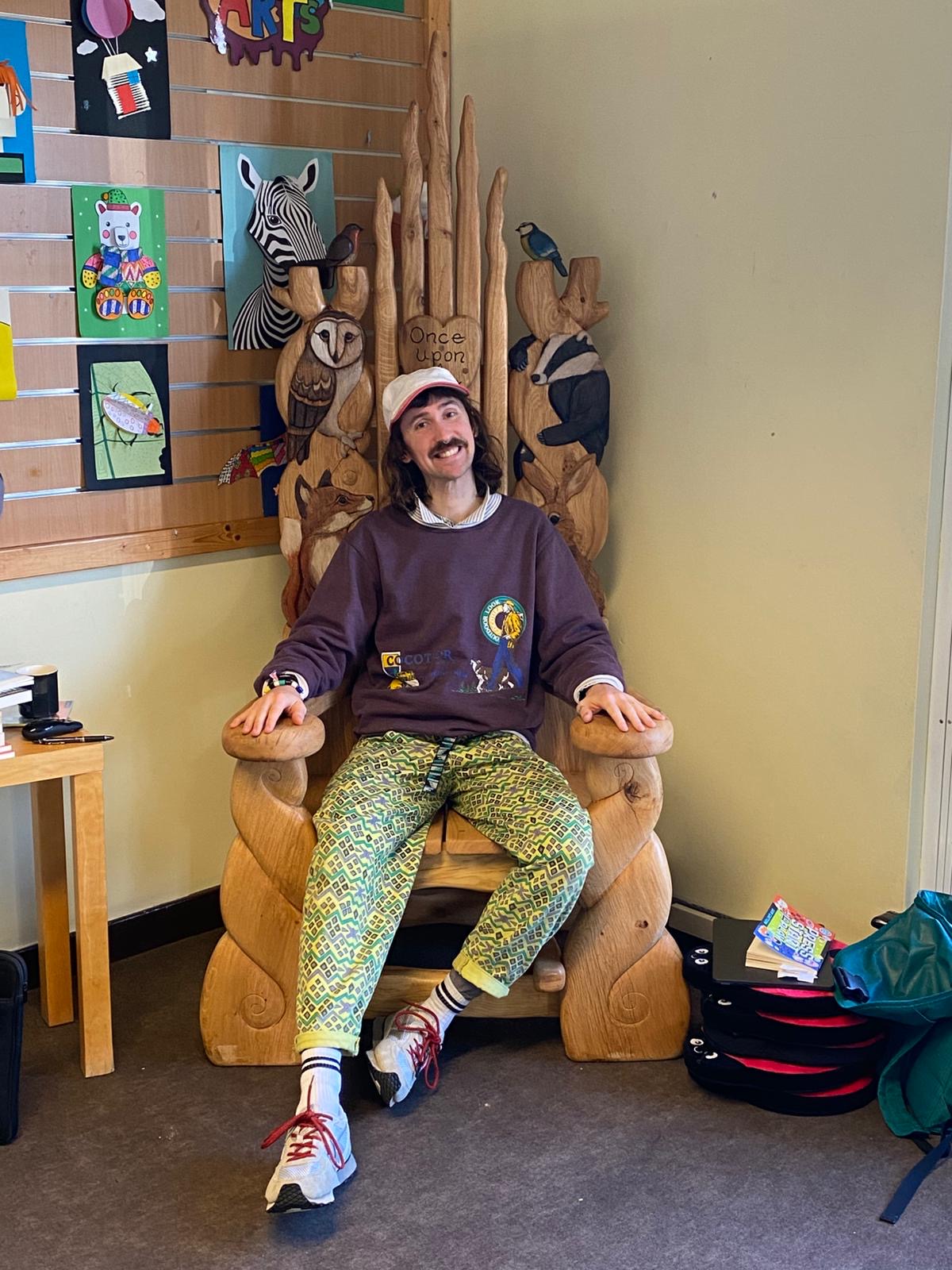 Author Sam Gayton sitting in handcrafted wooden storytelling throne at library event, surrounded by children's artwork