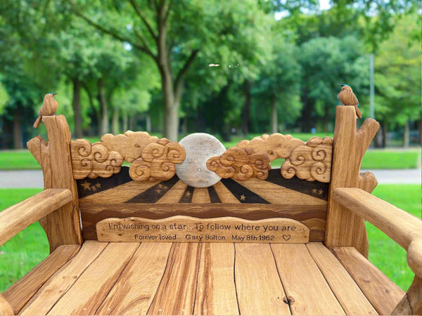Close-up of the wooden bench's backrest, showing intricate carvings of clouds and a moon, along with an engraved memorial message. The armrests feature carved birds, adding a whimsical touch to the bench