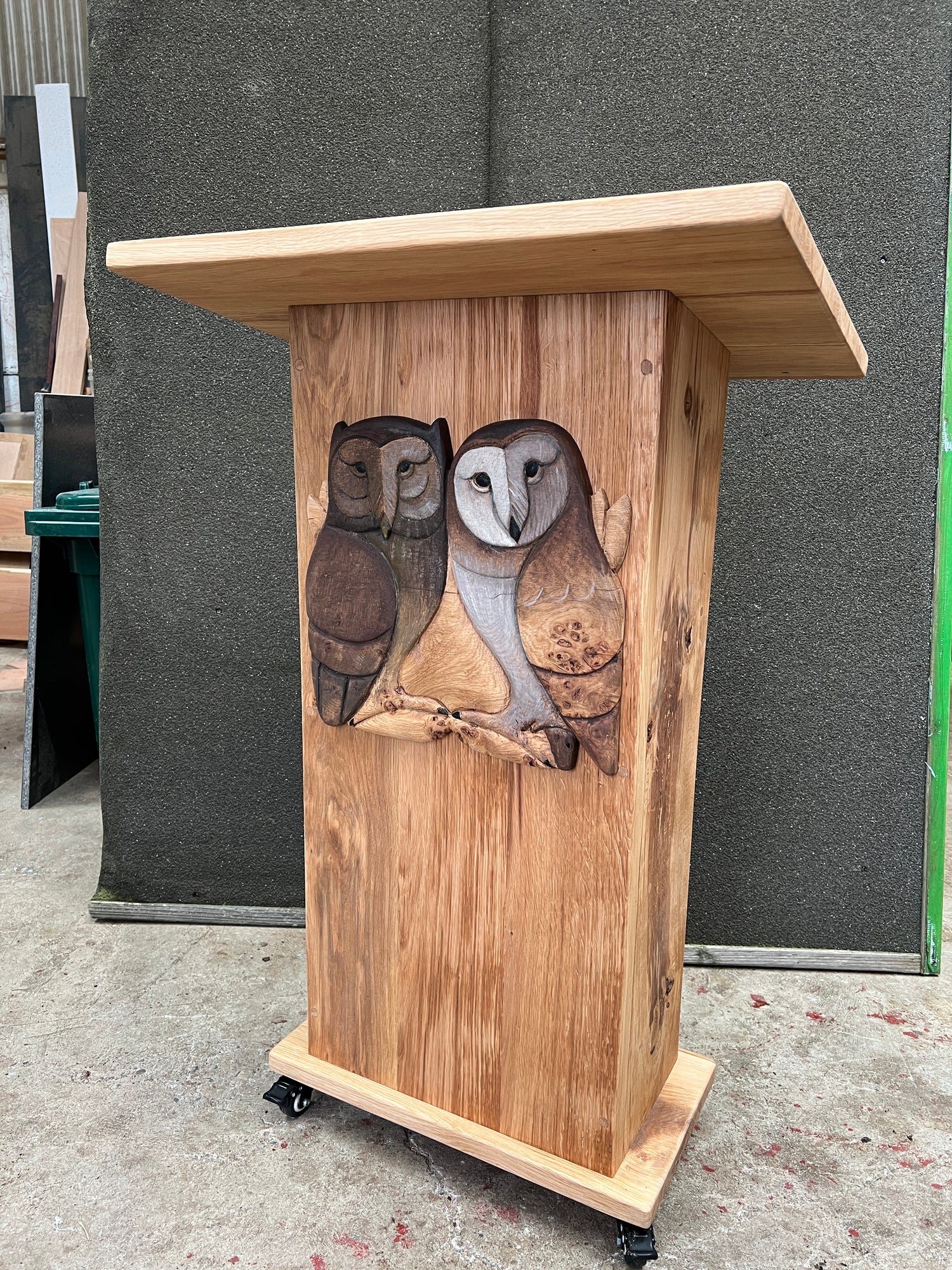 Front view of a handcrafted oak reading lectern with wheels, featuring two intricately carved owls on the front. The lectern is positioned in a workshop environment with various woodworking tools and materials visible in the background