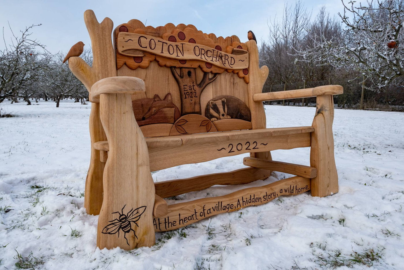 Banc en bois fabriqué à la main avec des sculptures complexes représentant un blaireau, un arbre et une abeille, avec l'inscription "Coton Orchard - Est. 1922" et "2022". Situé dans un verger enneigé