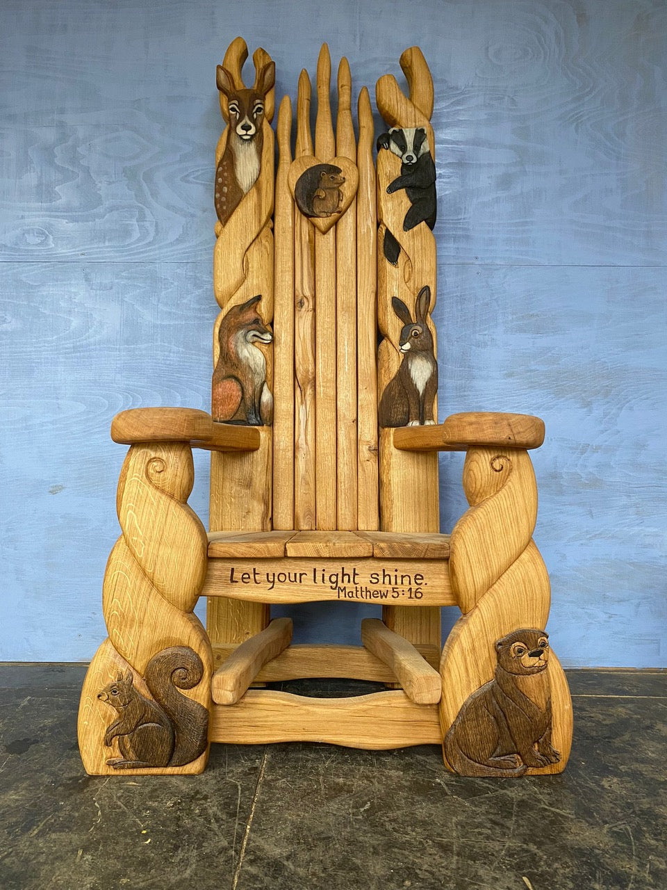 Handcrafted wooden storytelling chair featuring intricate animal carvings, including a deer, badger, fox, squirrel, and rabbit, with the words "Let your light shine. Matthew 5:16" engraved on the front, embodying a whimsical woodland theme