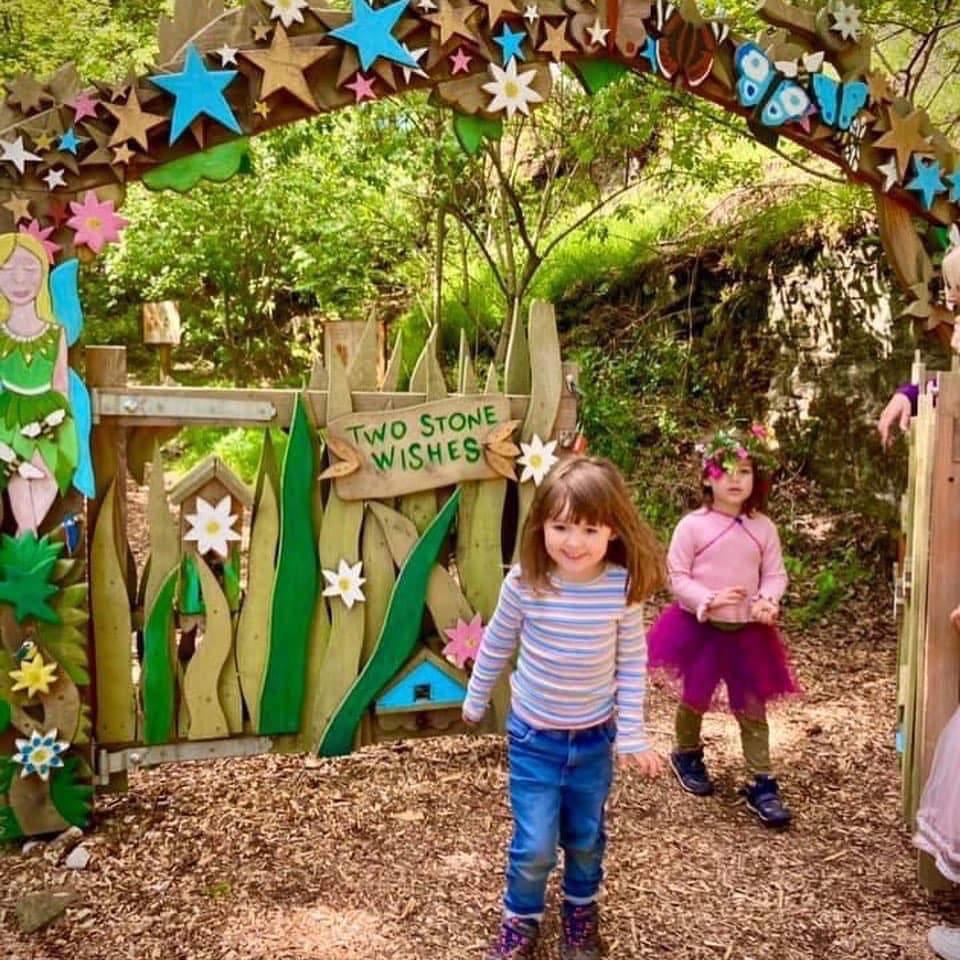 Children standing in front of a decorative wooden arch with fairy-themed decorations in a forest setting.