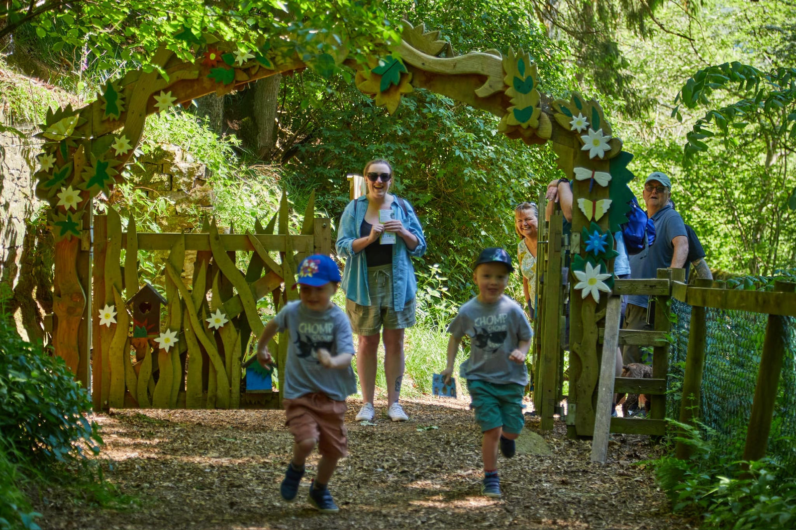 Children and adults walking through a wooden archway decorated with flowers and nature-themed elements in a forest setting.