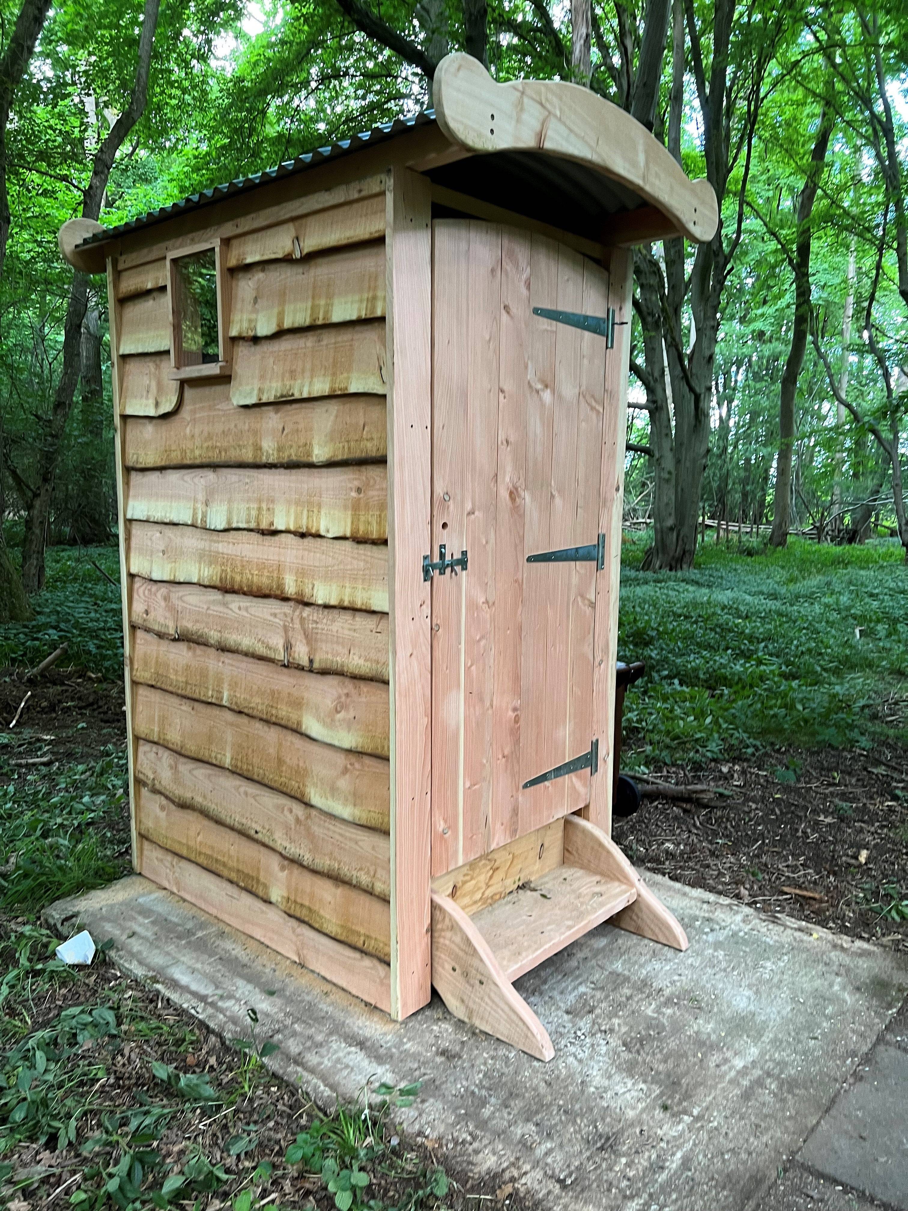 Wooden composting toilet with a rustic design, featuring a curved roof, side window, and front step, set in a natural forest environment