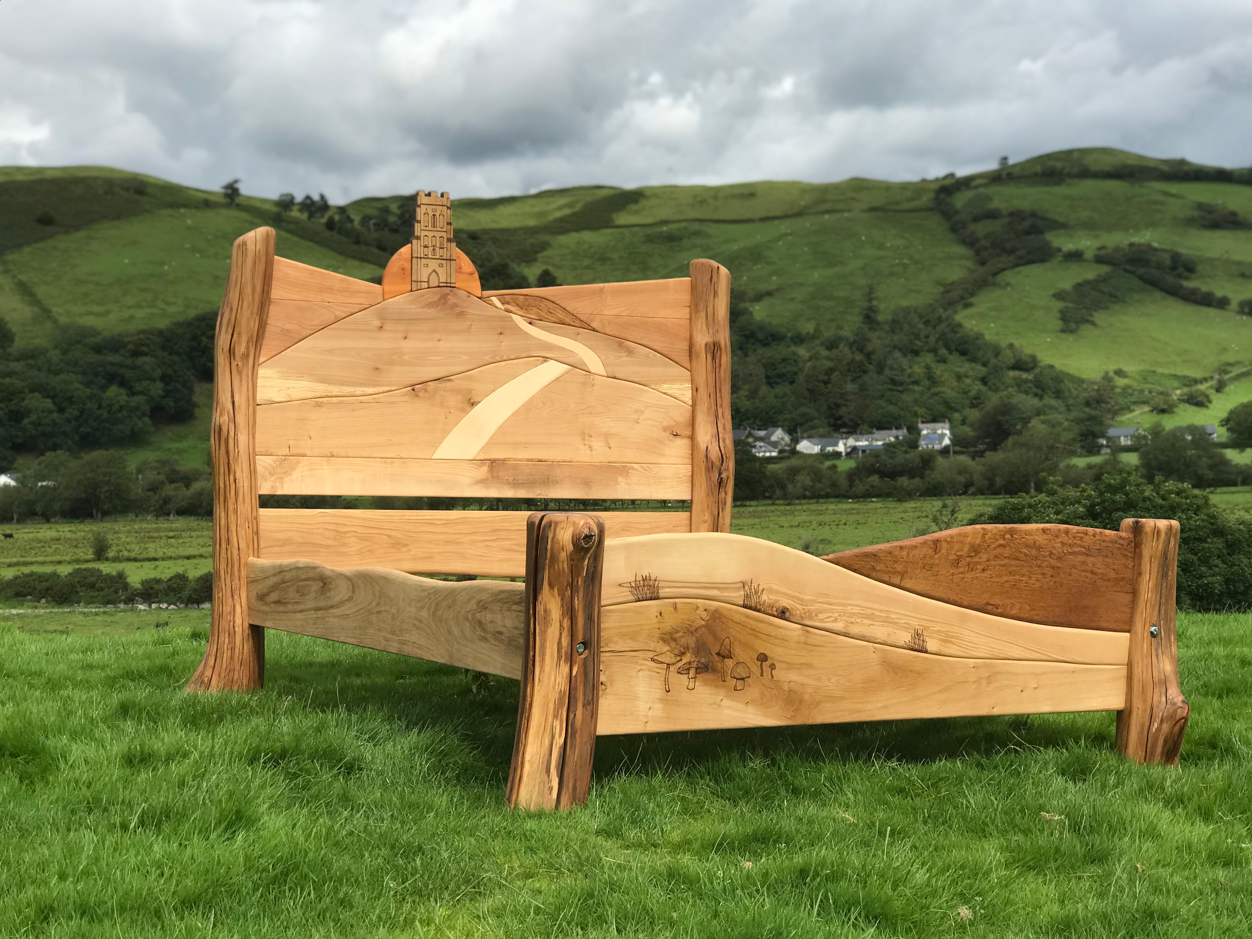 Glastonbury Tor bed with carved sunrise and path design, set against a rural landscape with green hills