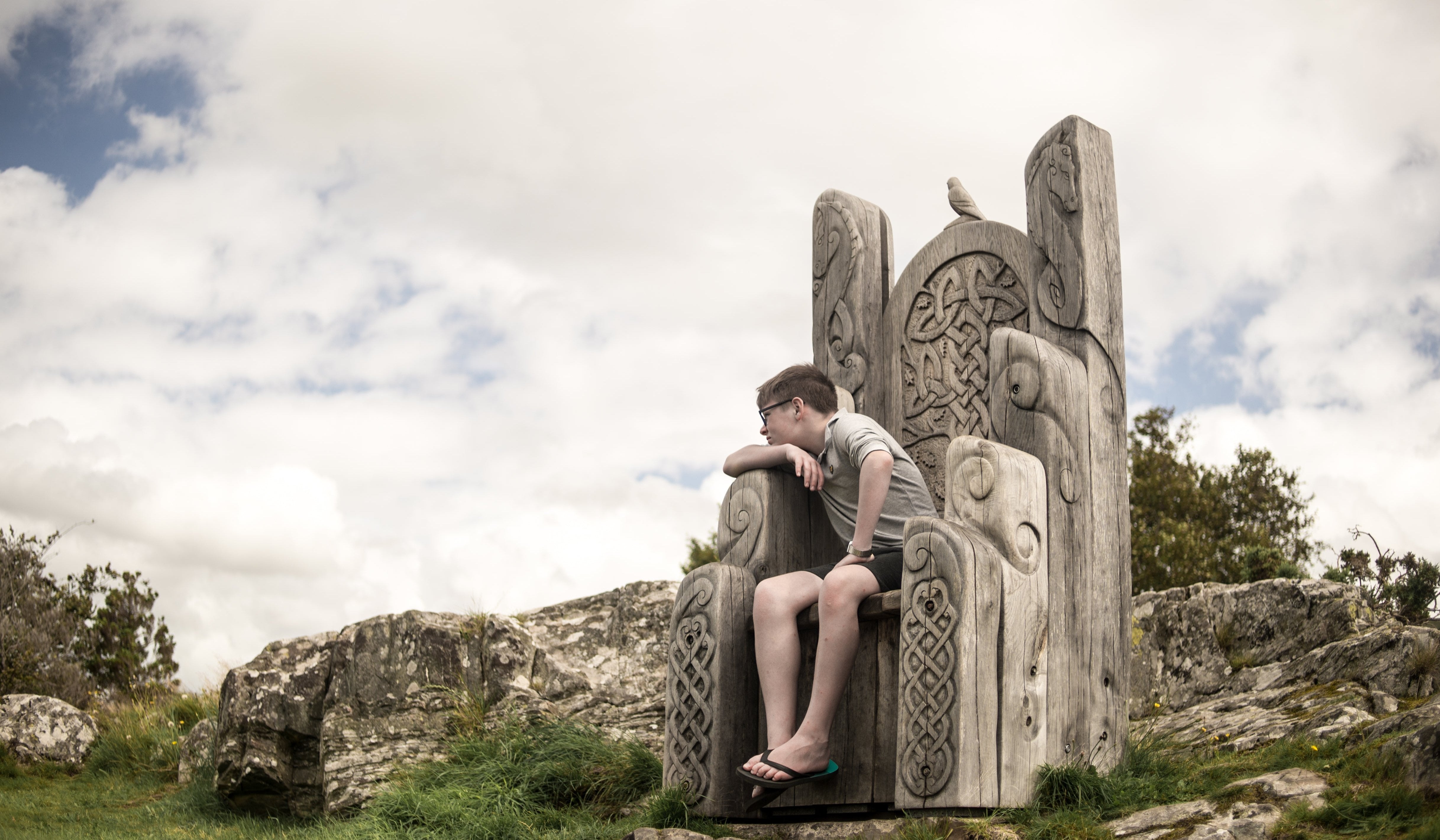 Jeune personne assise sur une grande chaise en bois sculptée de motifs celtiques, en plein air, sur fond de ciel nuageux.