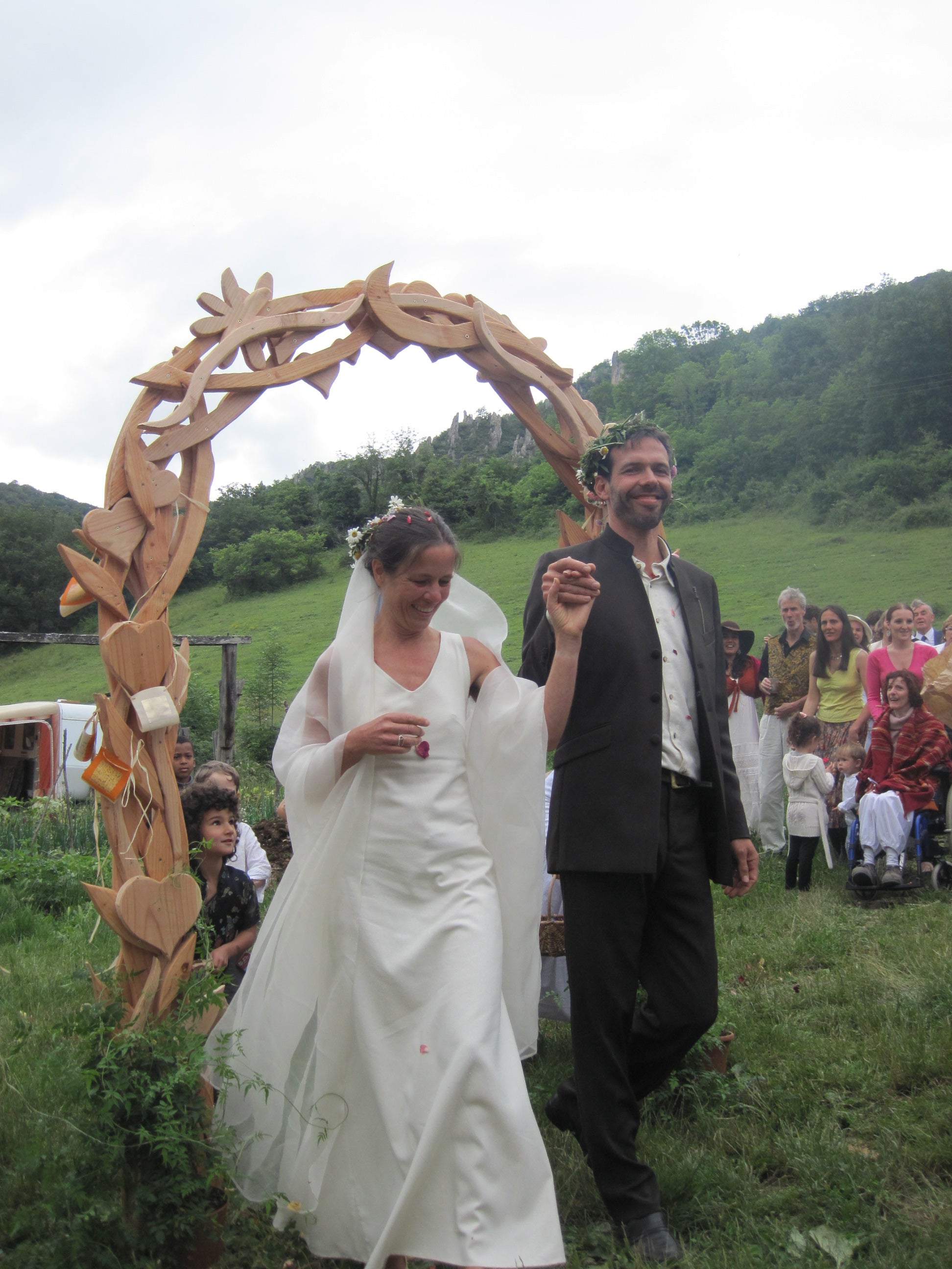 Two people in wedding attire standing under a wooden arch with a scenic background.