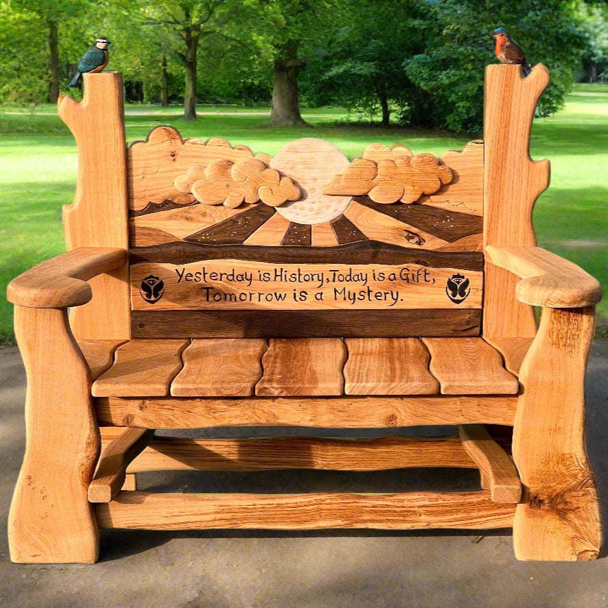 Handcrafted wooden memorial bench with a beautifully carved backrest featuring a sunrise, clouds, and a full moon. The bench is engraved with the quote: ‘Yesterday is History, Today is a Gift, Tomorrow is a Mystery.’ Two sculpted birds, a robin and a blue tit, perch on the rustic armrests, adding a natural, symbolic touch. The bench is set in a lush green park, creating a peaceful and reflective atmosphere