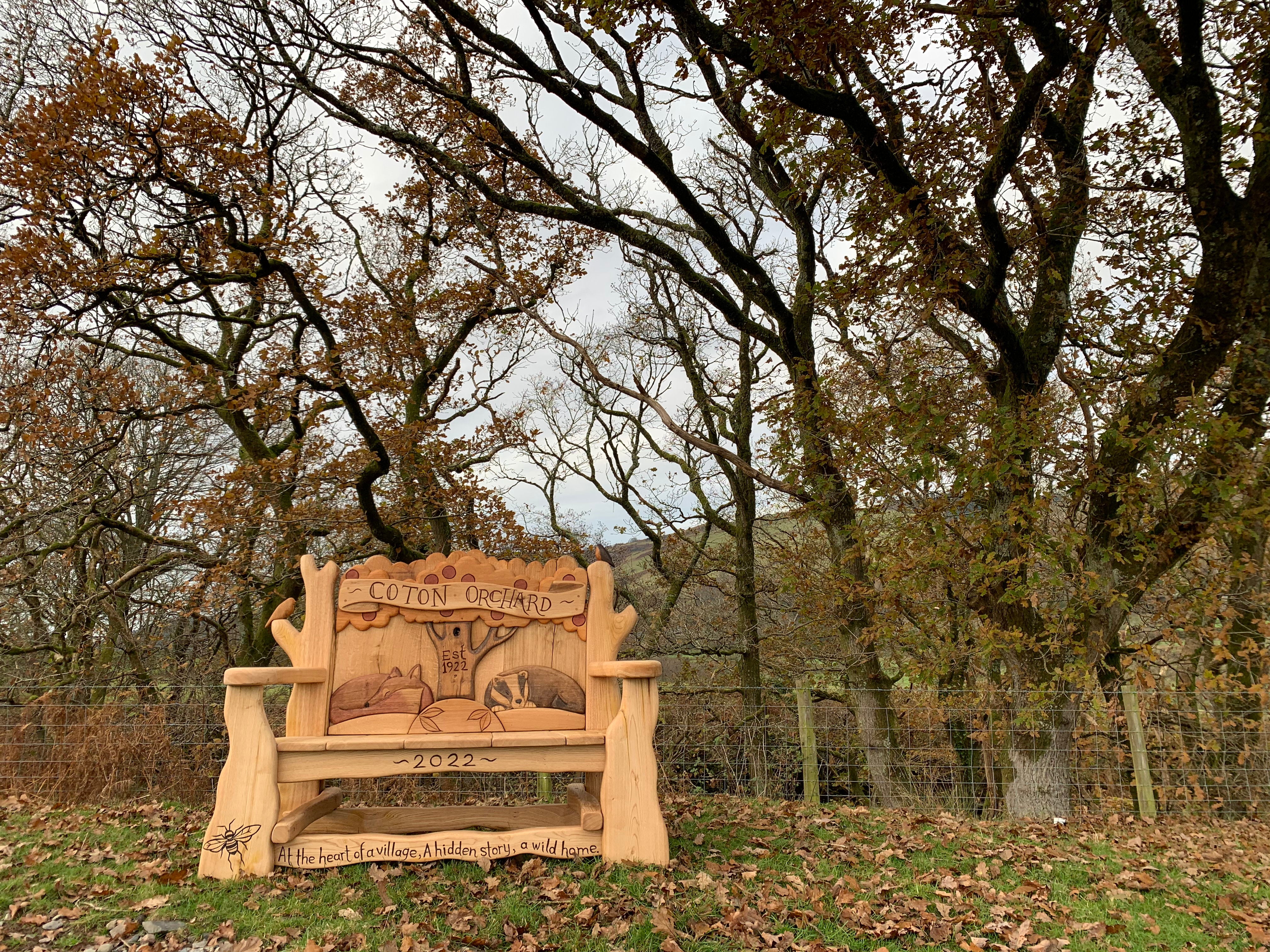 Coton Orchard Bench set in a scenic woodland, featuring detailed wildlife carvings of a fox, bee, and tree, celebrating heritage with the engraved inscription "At the heart of a village, A hidden story, a wild home