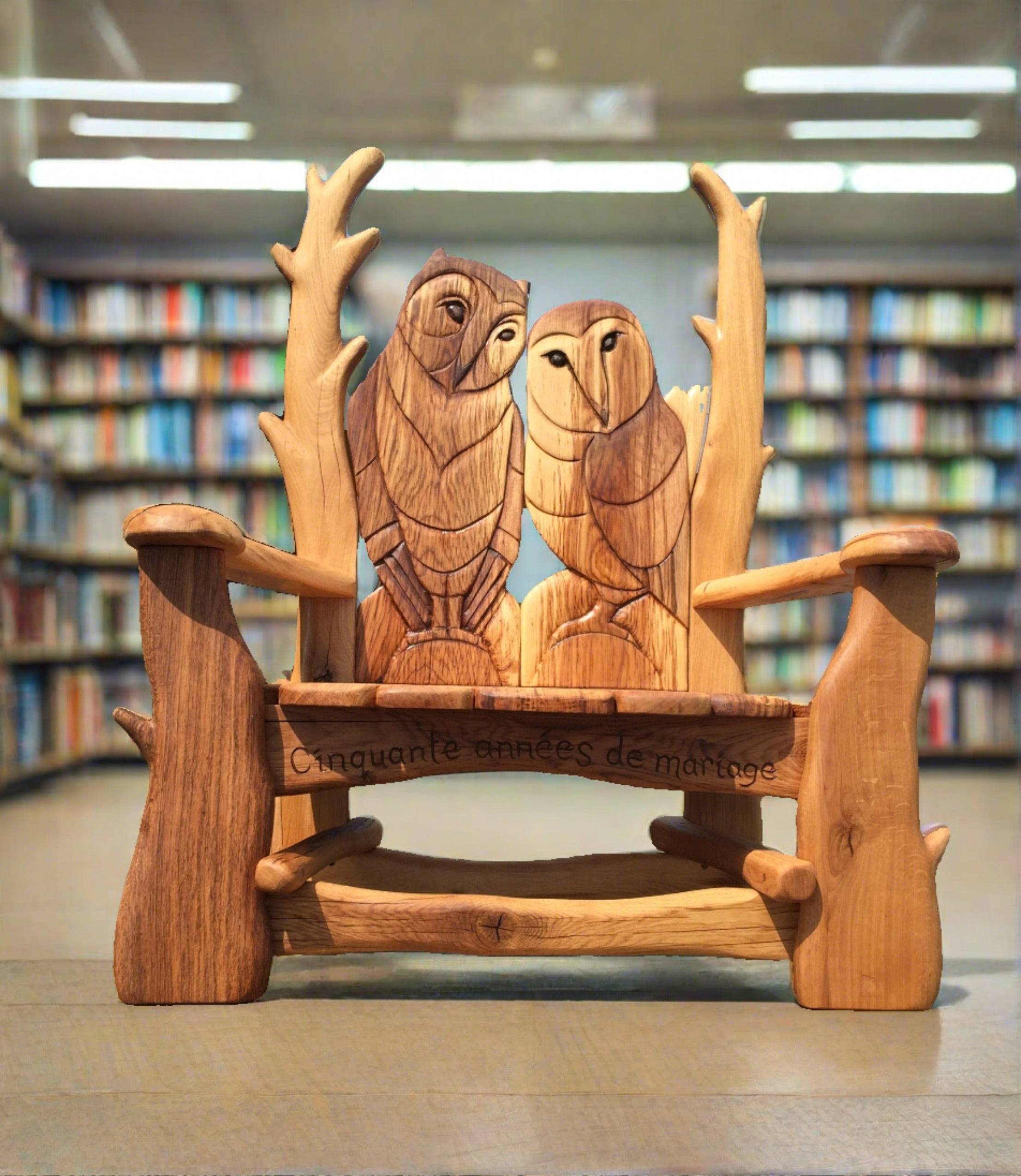 Handcrafted wooden bench featuring two intricately carved owls on the backrest. The bench includes an inscription 'Cinquante années de mariage' to commemorate fifty years of marriage, placed in a cozy library setting