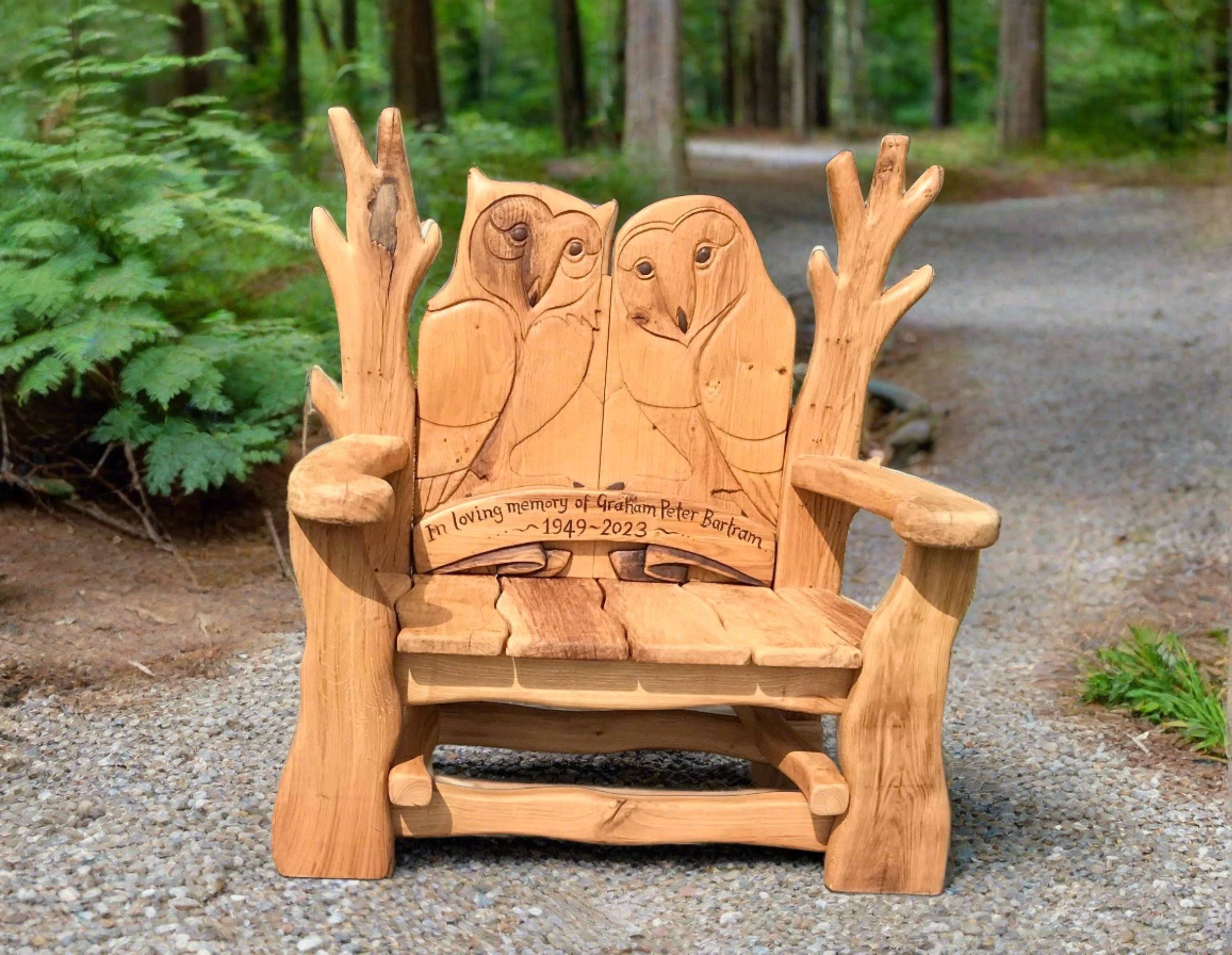 Handgefertigte Gedenkbank aus Holz mit zwei detaillierten Eulenschnitzereien auf der Rückenlehne. Die Bank ist mit der Gravur "In loving memory of Graham Peter Bartram 1949-2023" versehen und steht an einem Waldweg, umgeben von Grün.