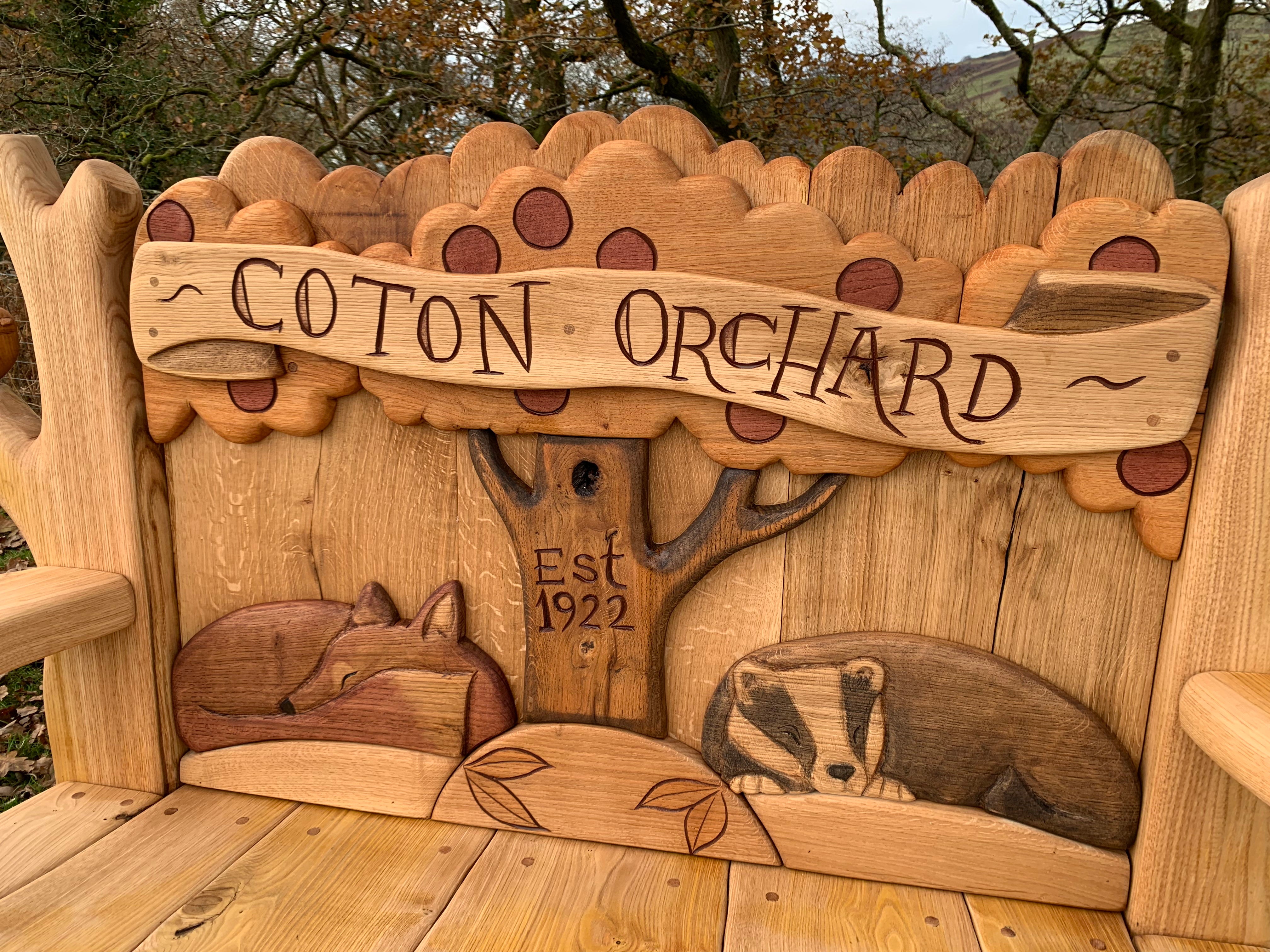 Close-up of the Coton Orchard Bench carvings featuring a fox, a badger, and a tree with the inscription "Est 1922," highlighting the intricate handcrafted details.