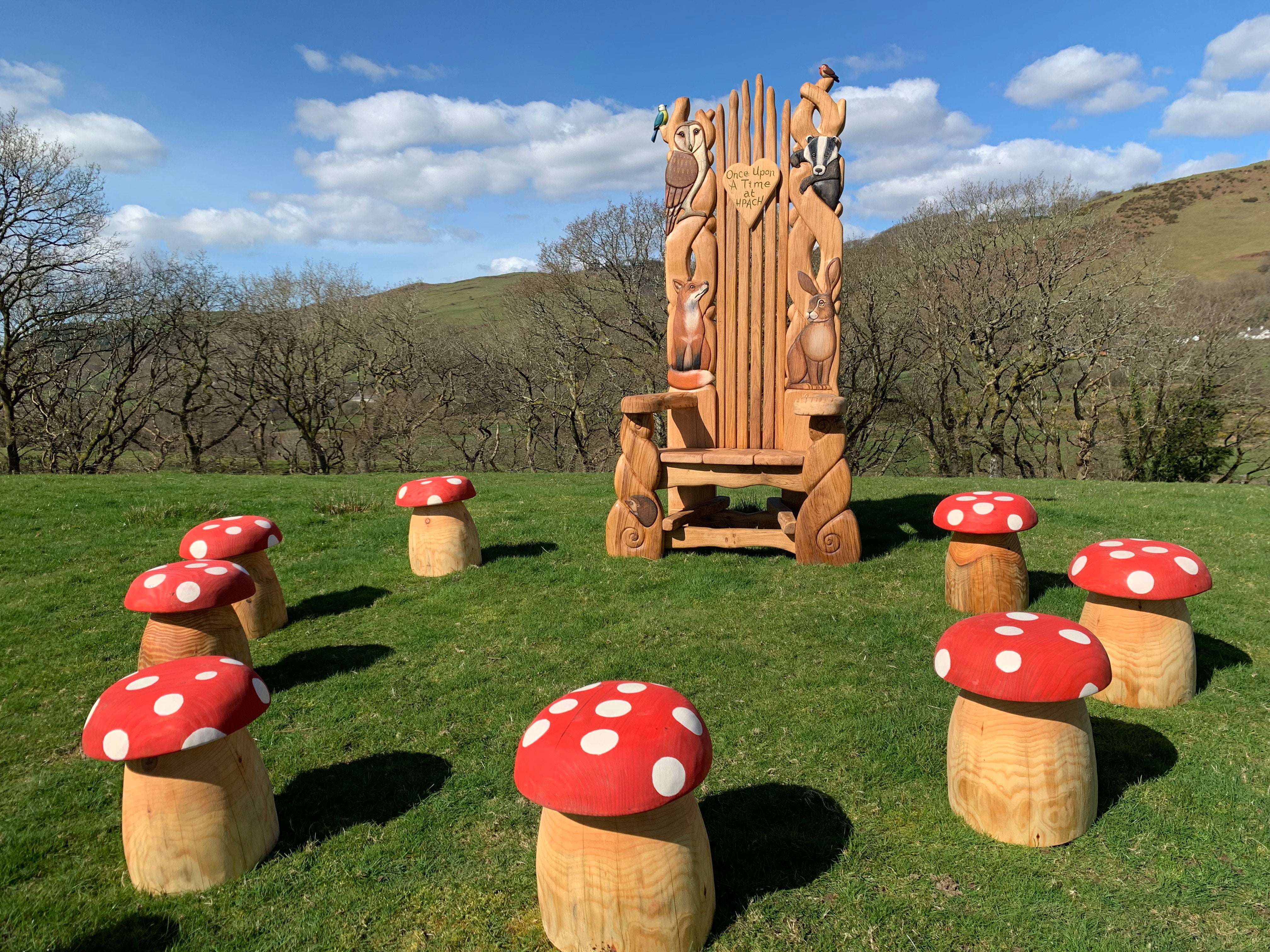 Skurriler Erzählstuhl, umgeben von Pilzhockern, handgeschnitzt mit Waldtieren, vor der malerischen Kulisse sanfter Hügel - perfekt, um die Fantasie anzuregen und Geschichten in der Natur zu erzählen