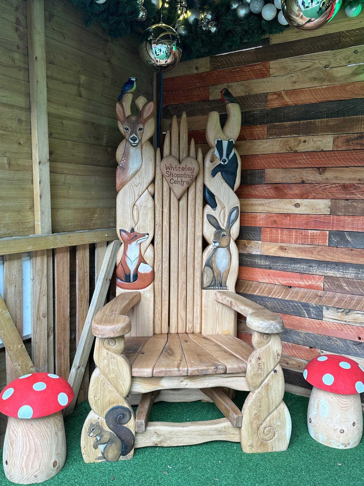 Woodland Creatures Storytelling Chair - Whiteley Shopping CentreA beautifully crafted wooden storytelling chair featuring intricate carvings of woodland creatures including a deer, fox, badger, squirrel, and rabbit, with vibrant painted details. The chair is engraved with "Whiteley Shopping Centre" on a heart-shaped plaque and is surrounded by festive decor, including red and white mushroom ornaments