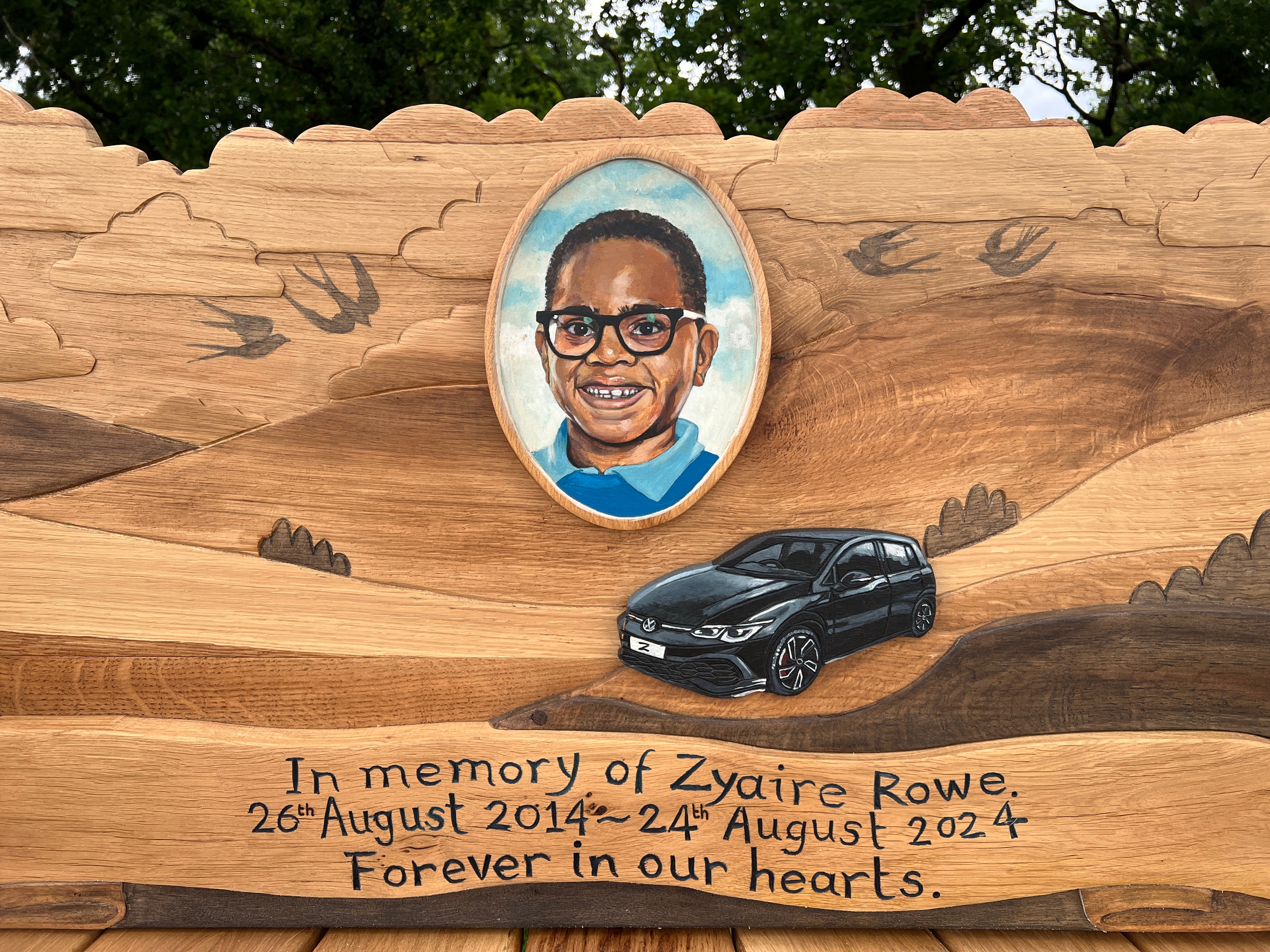 A handcrafted wooden memorial bench featuring a painted portrait of Zyaire Rowe in an oval frame, carved swallows, and a black car in relief, with engraved text reading “Forever in our hearts.”

