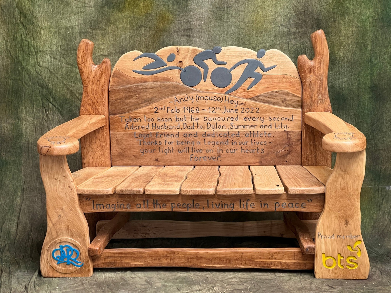 Detail of engraved memorial message for Andy “Mouse” Hey on a carved wooden bench, with smooth oak grain and curved backrest.
