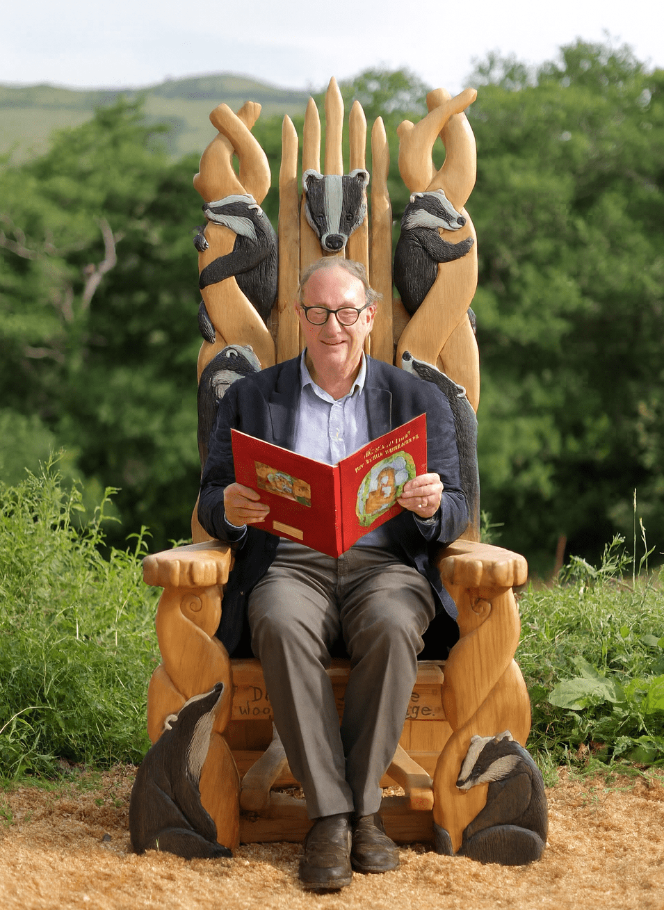 Man sitting on a throne made of badger sculptures, reading a book outdoors.
