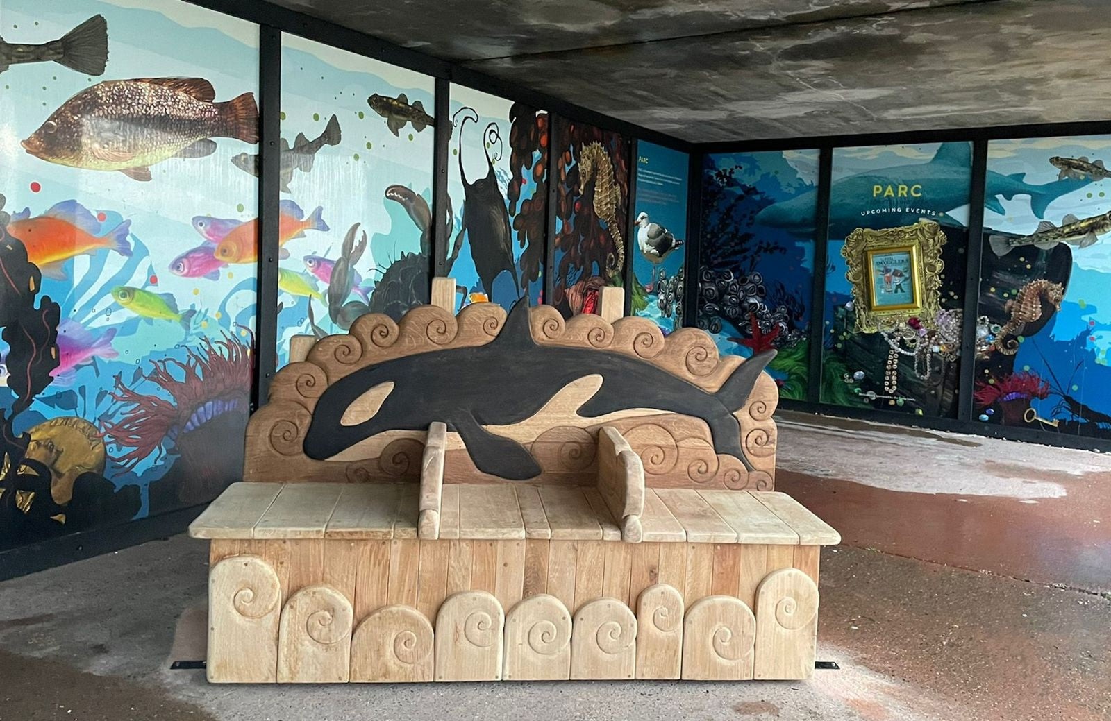 Wide view of oak orca memorial bench installed on Rottingdean seafront with marine mural backdrop