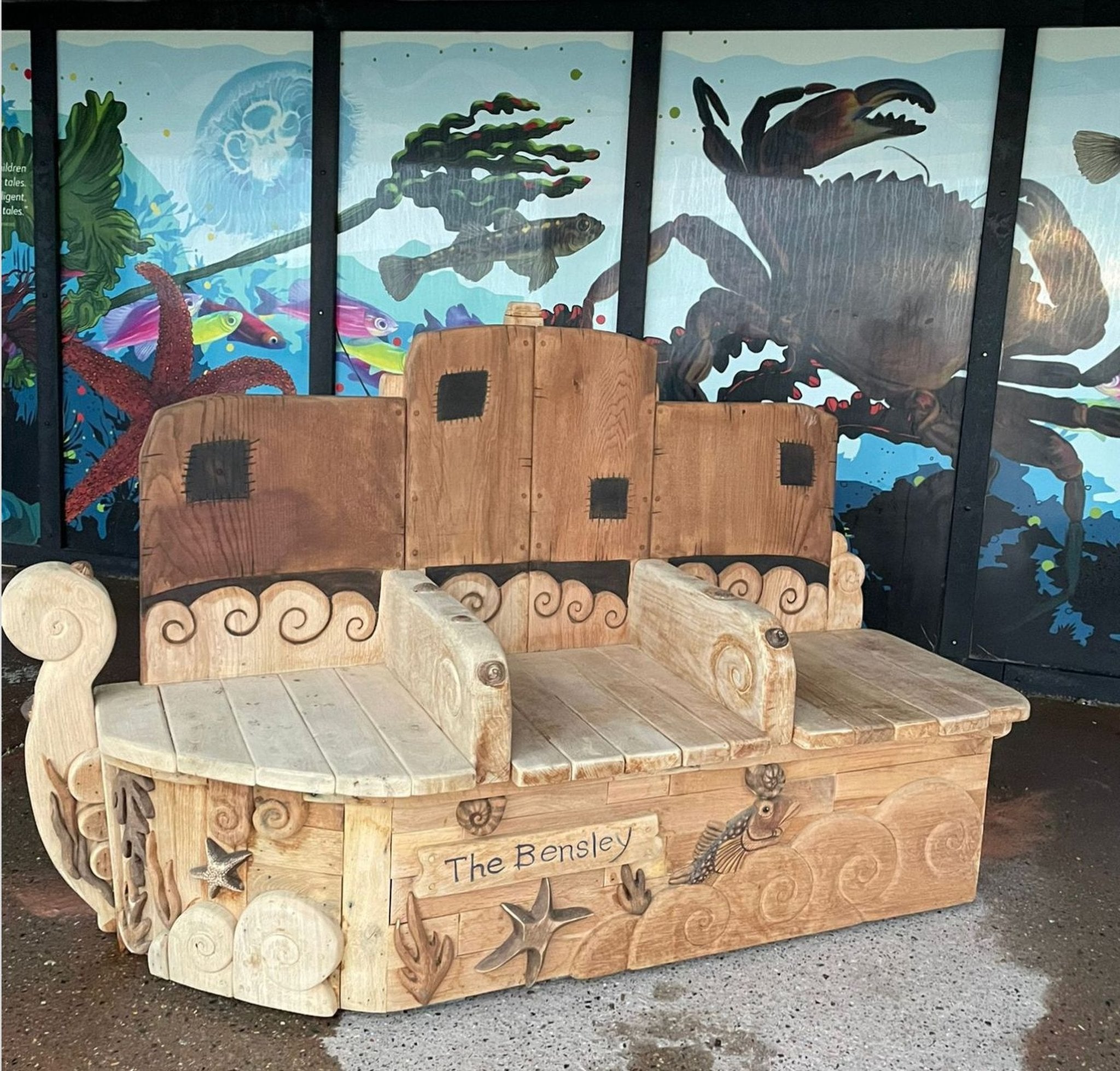 Wide view of The Bensley hand-carved oak boat memorial bench on Rottingdean seafront with marine mural backdrop