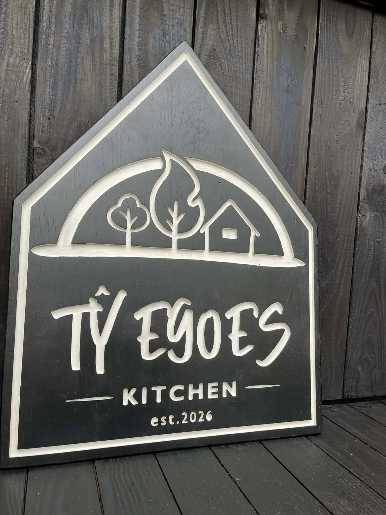 Angled view of custom engraved oak business sign with house silhouette shape and deep charcoal painted finish