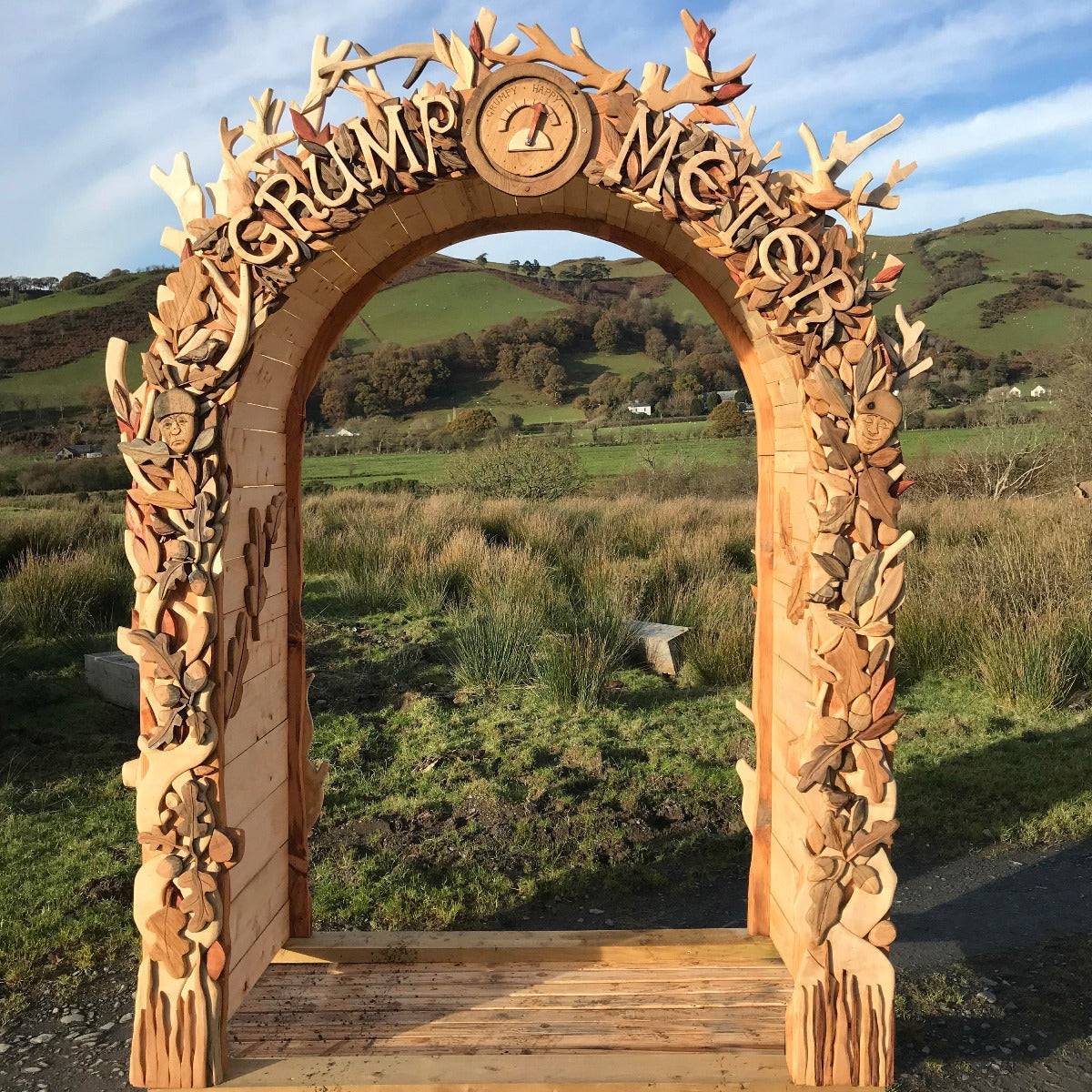 Hand-carved wooden arch titled 'Grump Meter,' created for Lapland Festival. The arch features intricate carvings of leaves, branches, and whimsical faces, set against a scenic backdrop of rolling green hills