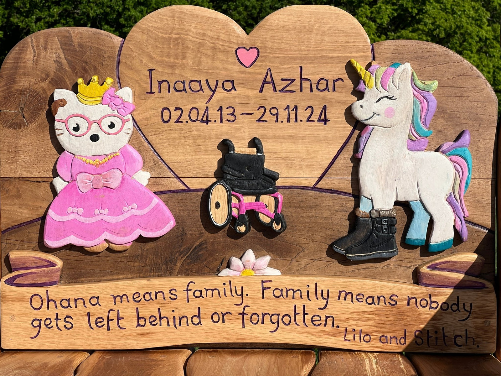 Close-up of personalised children’s memorial bench backrest with name, dates, and colourful carvings of a princess cat, a wheelchair, and a unicorn, along with the quote “Ohana means family. Family means nobody gets left behind or forgotten.”