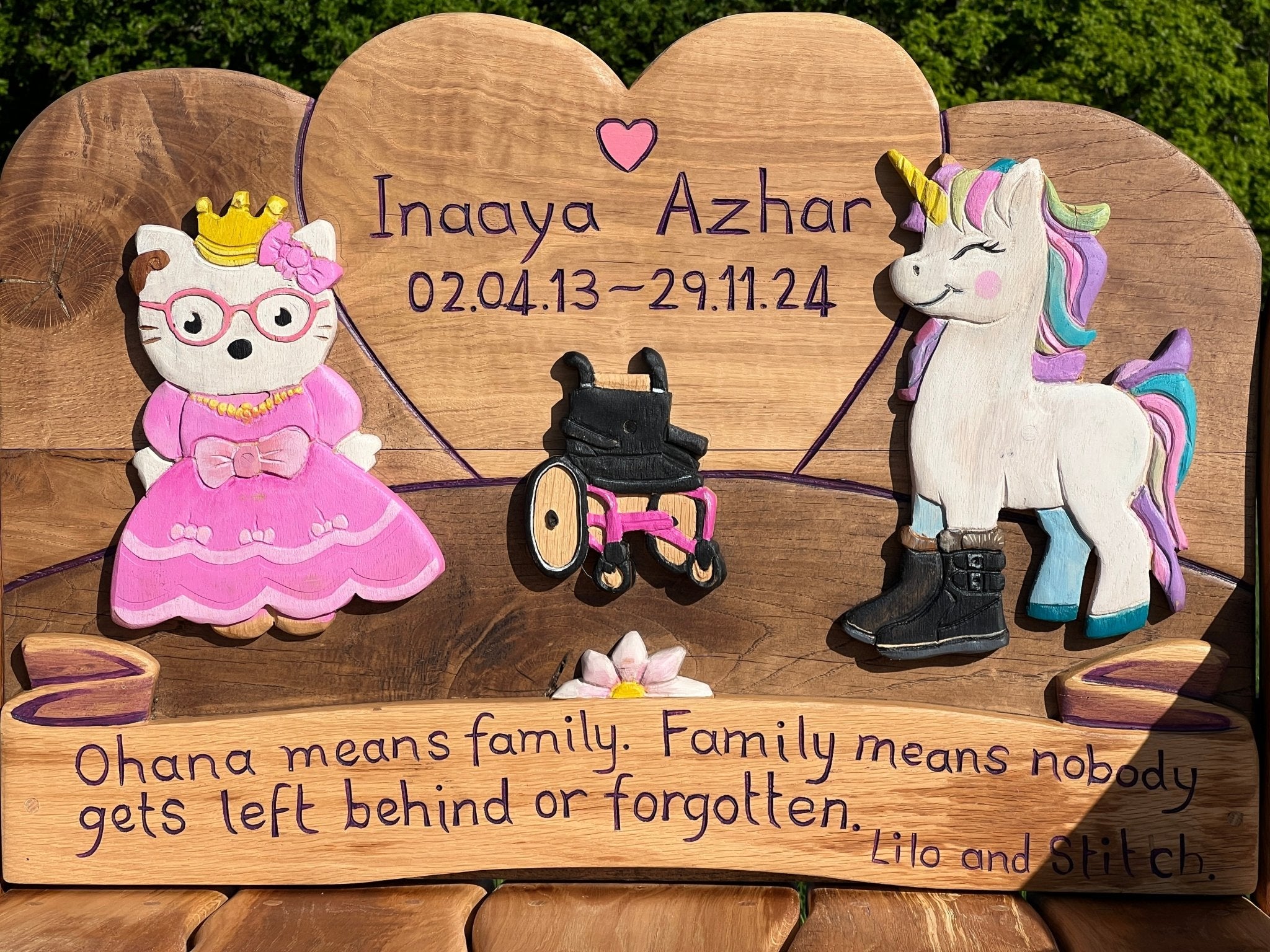 Close-up of personalised children’s memorial bench backrest with name, dates, and colourful carvings of a princess cat, a wheelchair, and a unicorn, along with the quote “Ohana means family. Family means nobody gets left behind or forgotten.”
