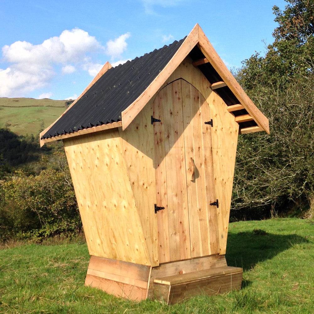 Stylish wooden Designer Composting Toilet with a unique slanted design, crafted from sustainable materials, featuring a pitched roof and rustic charm, set in a picturesque outdoor environment.