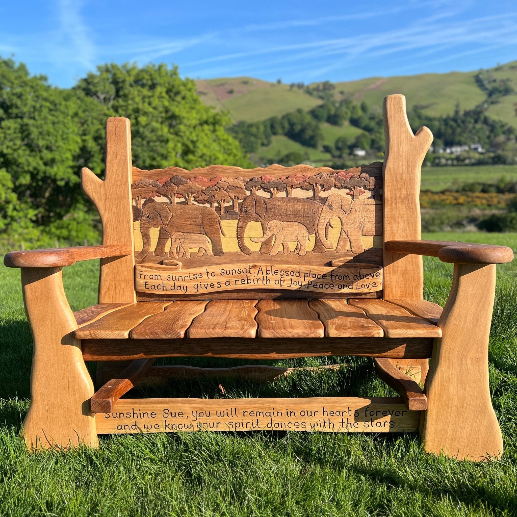 Hand-carved oak memorial bench with elephant family design and personalised engraved tribute text, set in a grassy landscape.