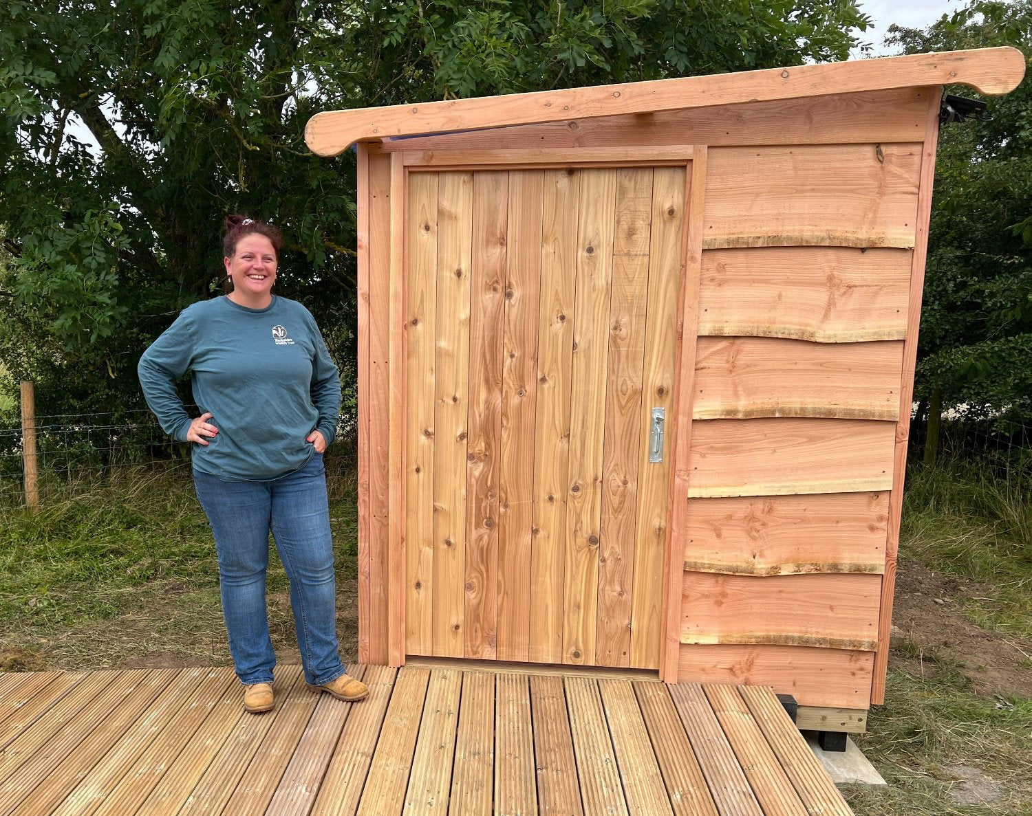 Woman standing beside a handcrafted disabled-access compost toilet installed for a church, featuring natural wood siding and accessible design elements