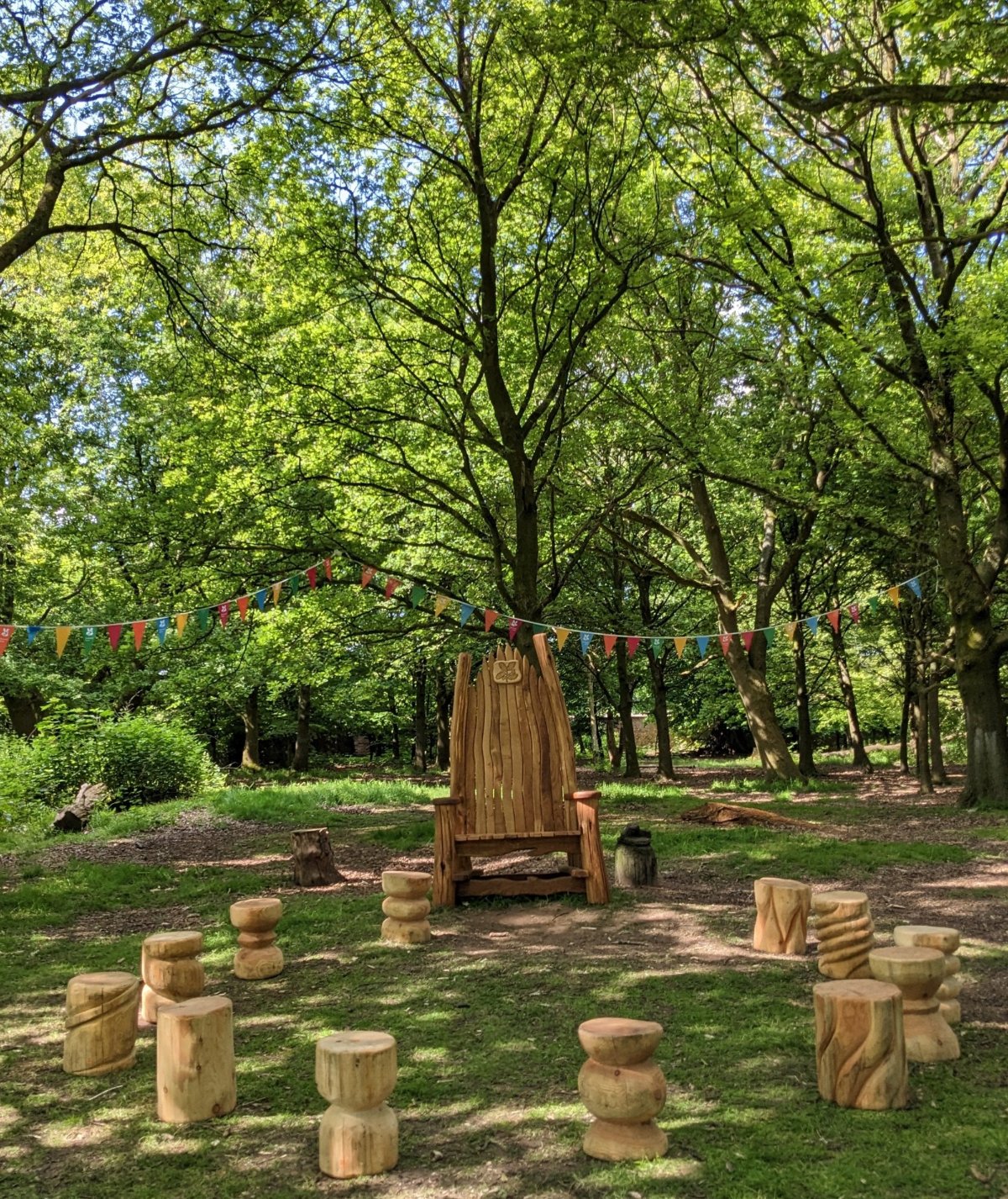 A beautifully carved wooden storytelling throne surrounded by wooden stools in a serene forest setting, adorned with colorful bunting and dappled sunlight filtering through the trees