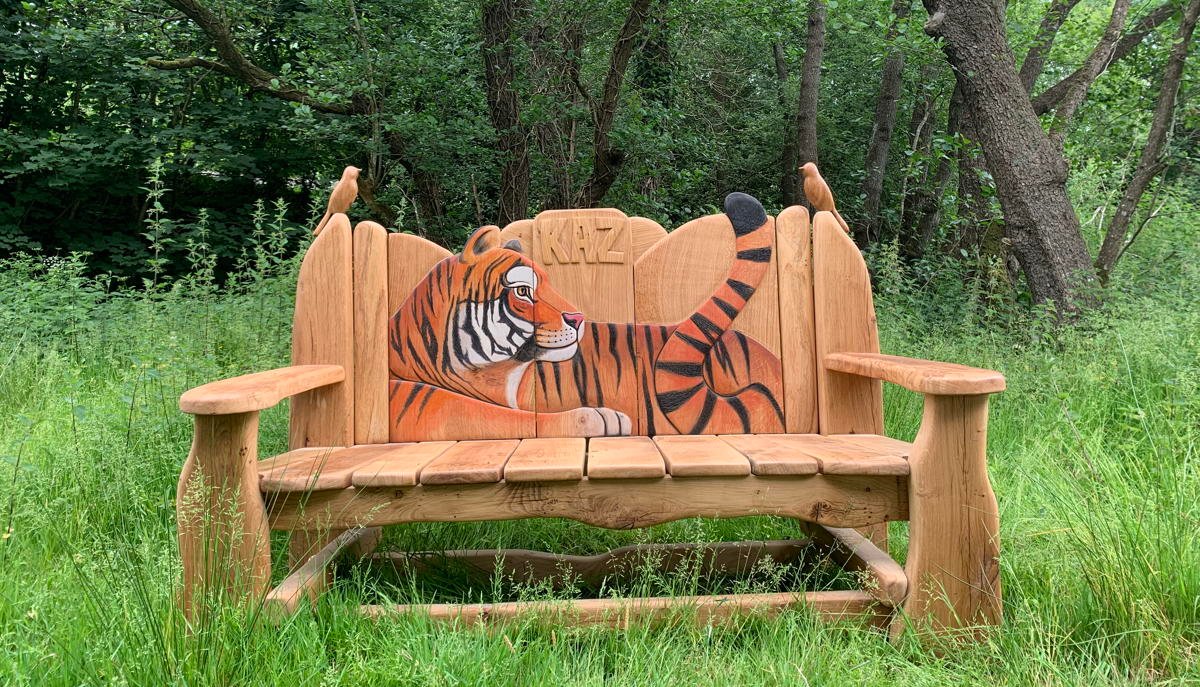 Handmade oak bench featuring a vibrant tiger carving, surrounded by natural forest greenery. Custom outdoor wooden bench by Free Range Designs