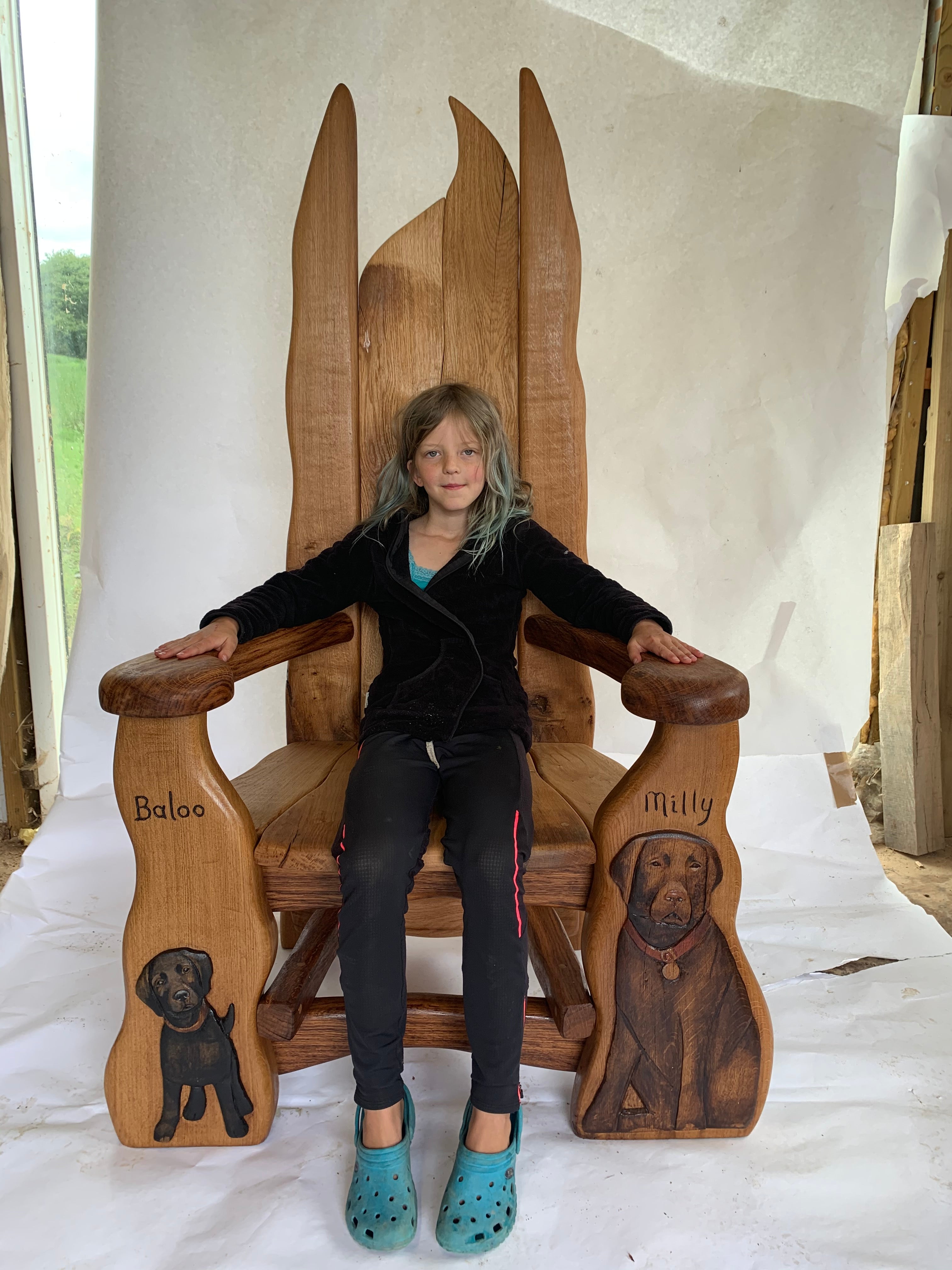 A beautifully handcrafted wooden chair featuring intricate carvings of dogs named Baloo and Milly, with a young girl seated, showcasing its artistic and functional design.